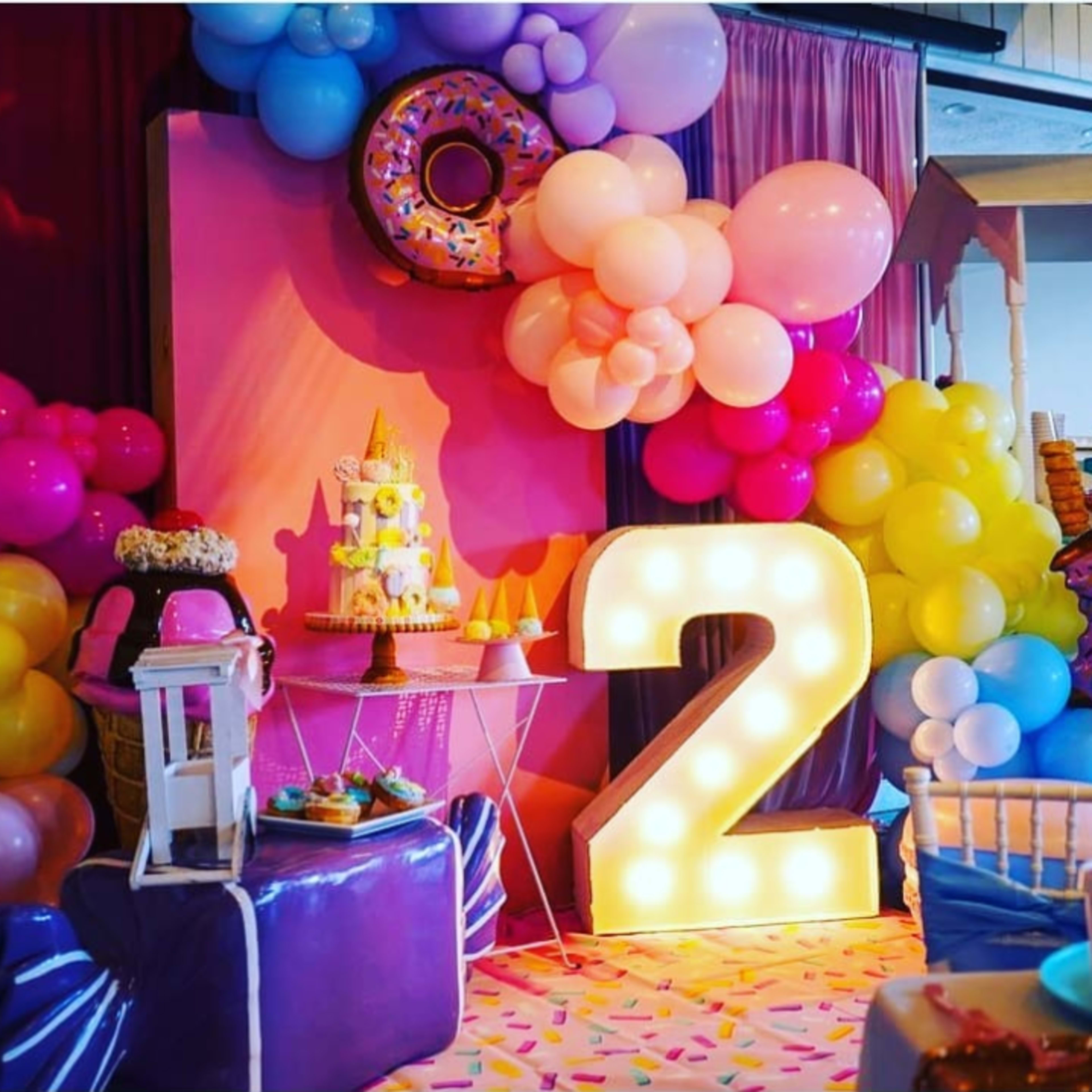 The scene features a colorful birthday party setup with balloons, a cake, and a large illuminated "2" decoration.