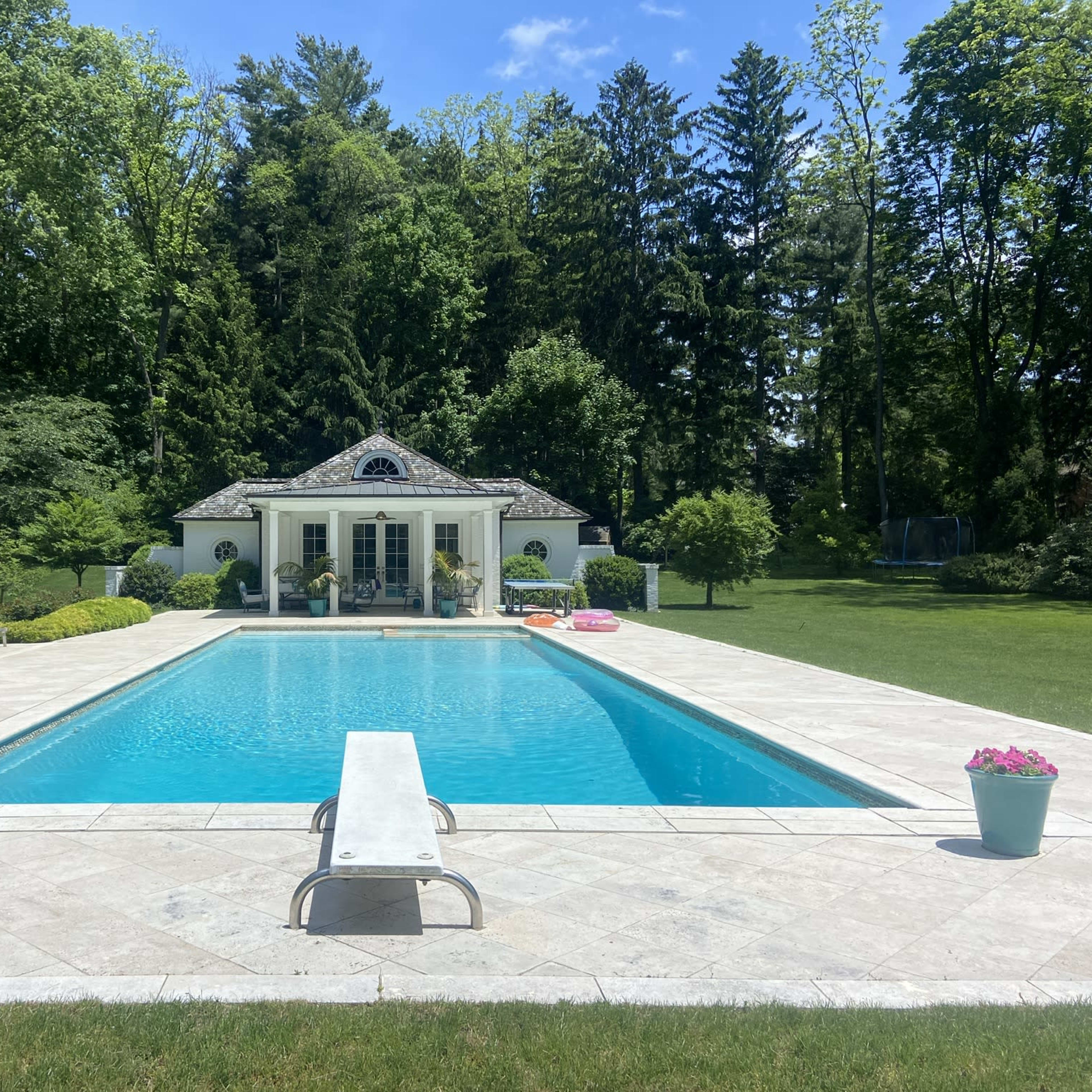 A rectangular swimming pool is surrounded by a manicured lawn, lounge chairs, and potted plants, with a white gazebo visible in the background against a backdrop of trees.