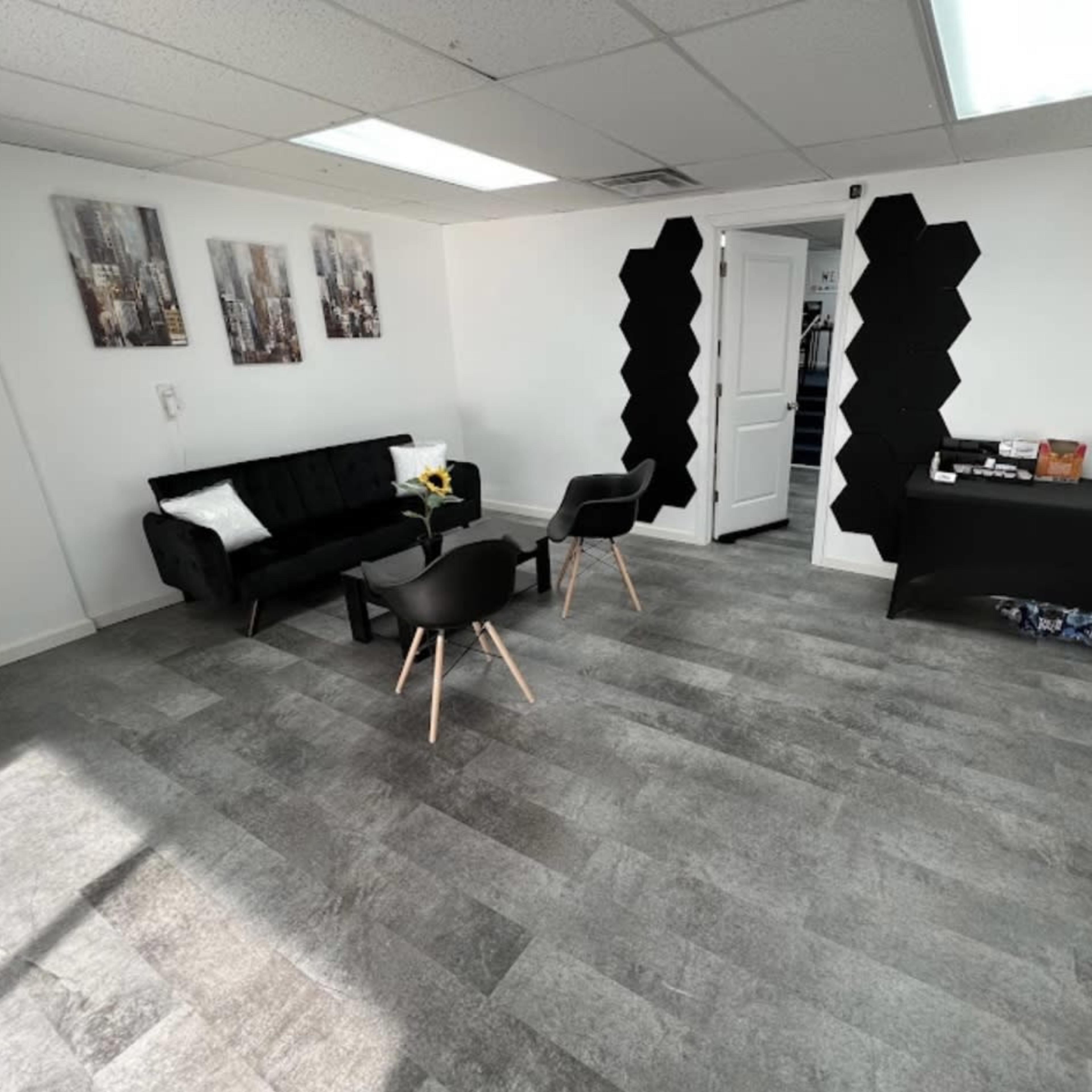 The room features a black couch, two black chairs, a side table, and a dark reception desk against a light-colored wall with abstract wall art.