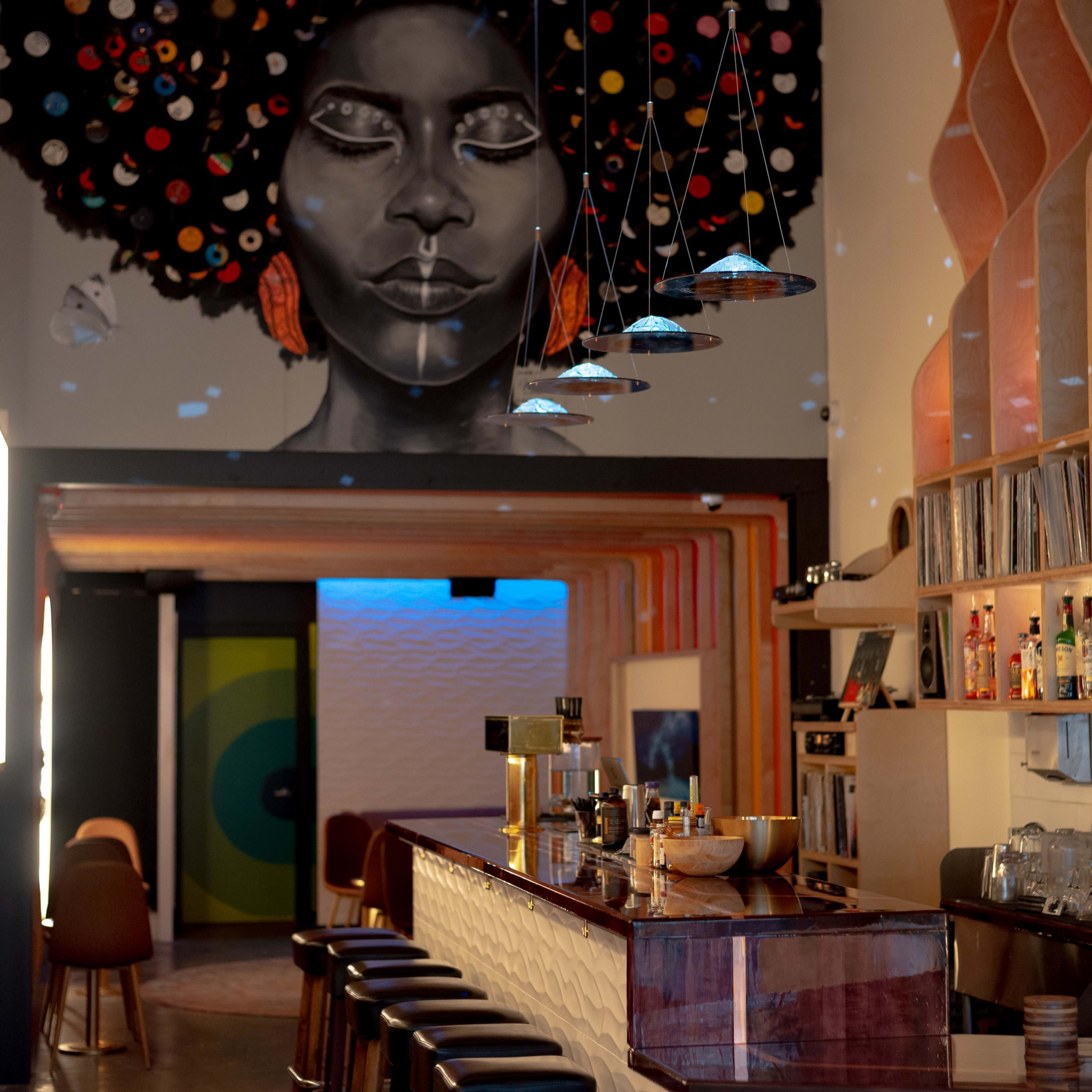 A spacious bar with a vibrant mural of a woman's face on the wall, surrounded by warm wooden accents and a well-stocked shelf of beverages.
