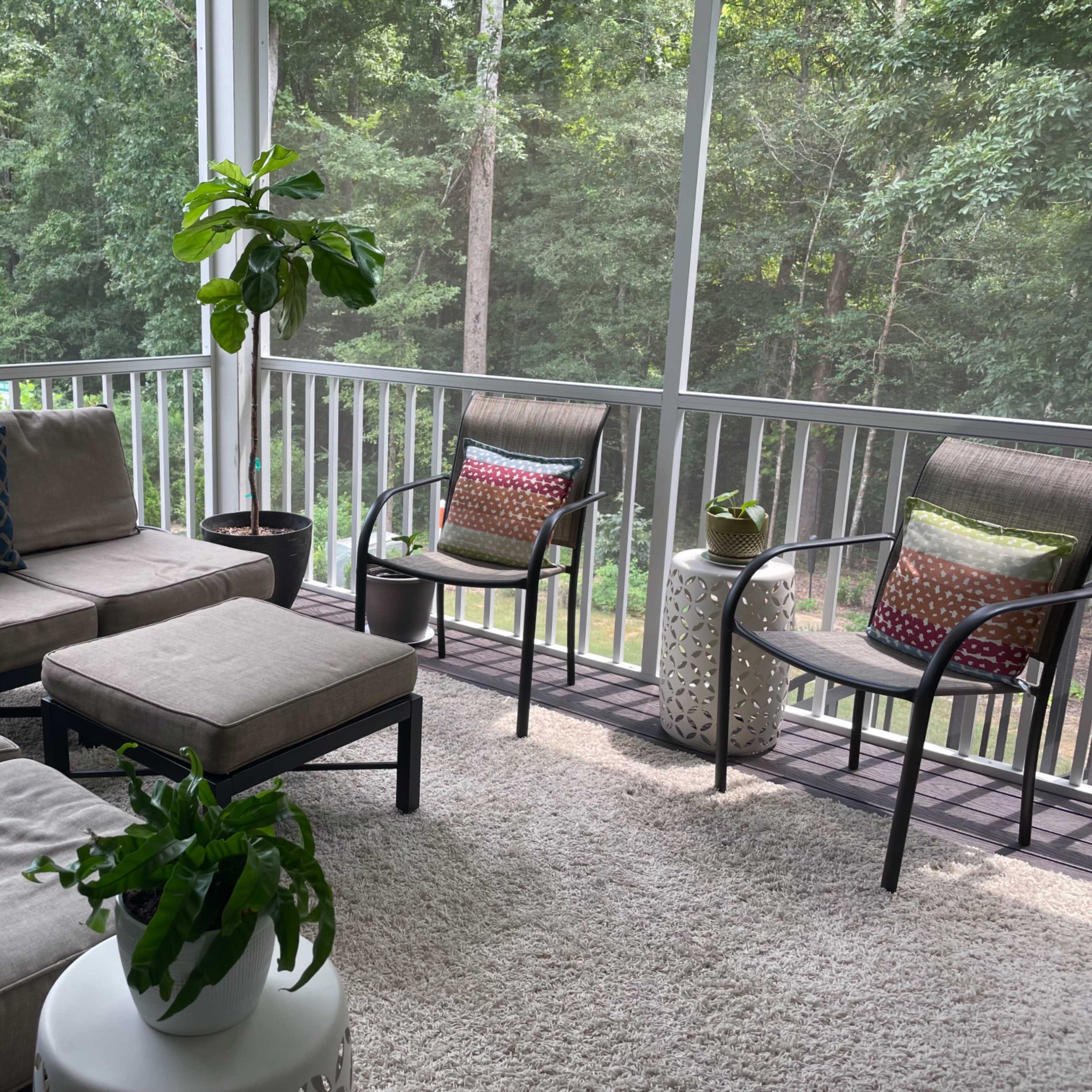 A screened-in porch features a sectional sofa, two chairs with colorful cushions, a coffee table, and potted plants, set against a backdrop of dense greenery.