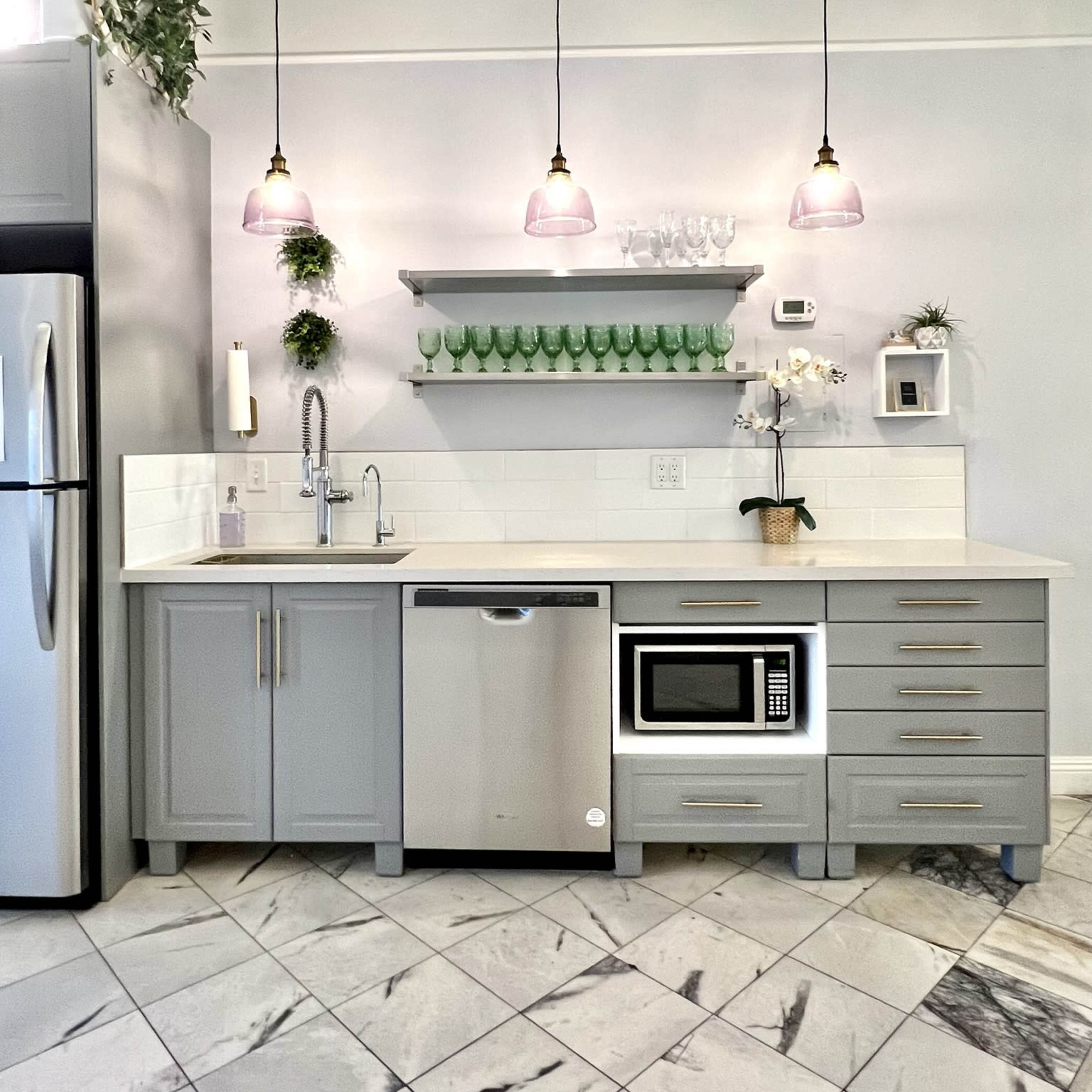 A modern kitchen features gray cabinetry, a stainless steel refrigerator, a microwave, and pendant lighting above a white countertop with green dishware on a shelf.