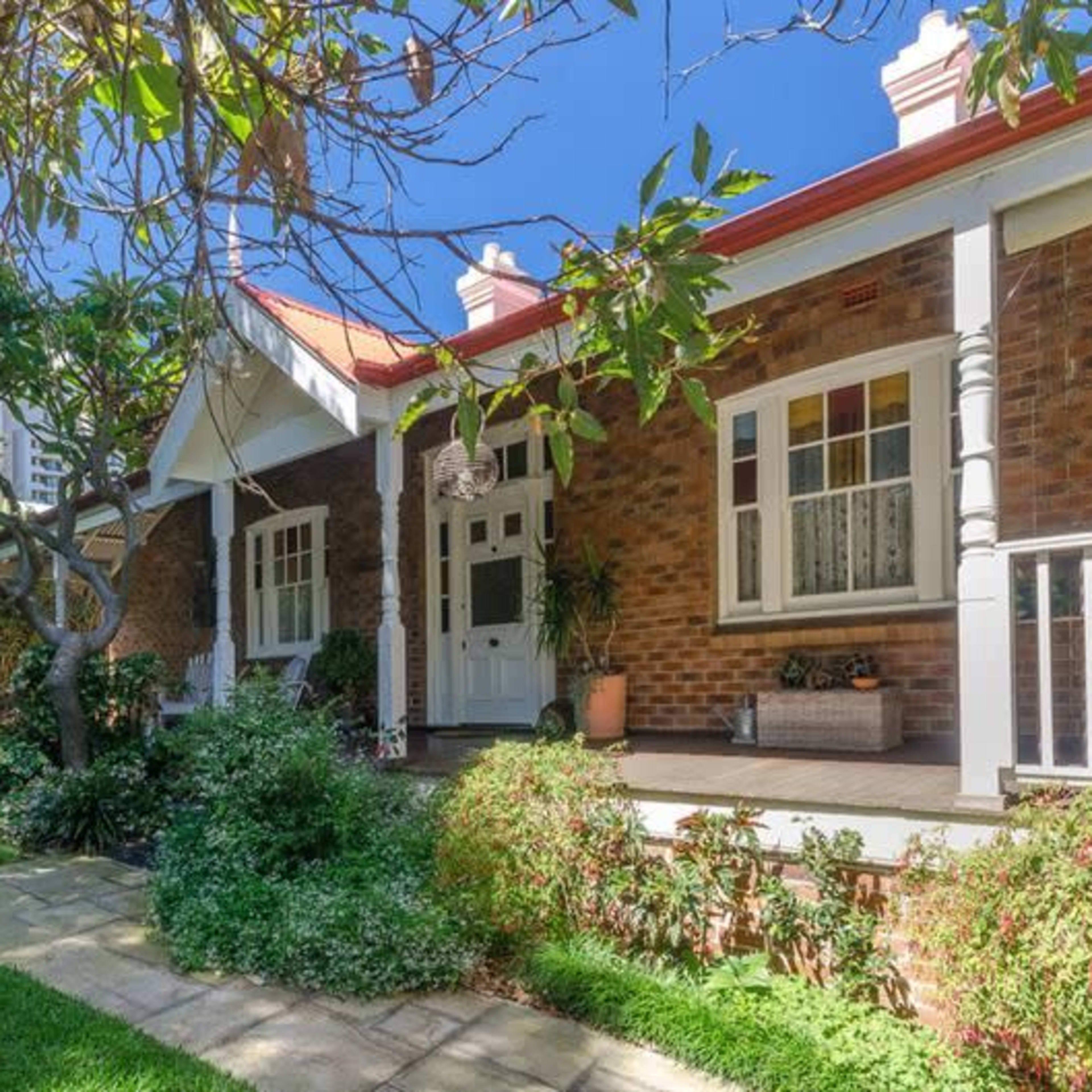 A brick house with a front porch and garden, featuring a pathway, trees, and lush greenery.