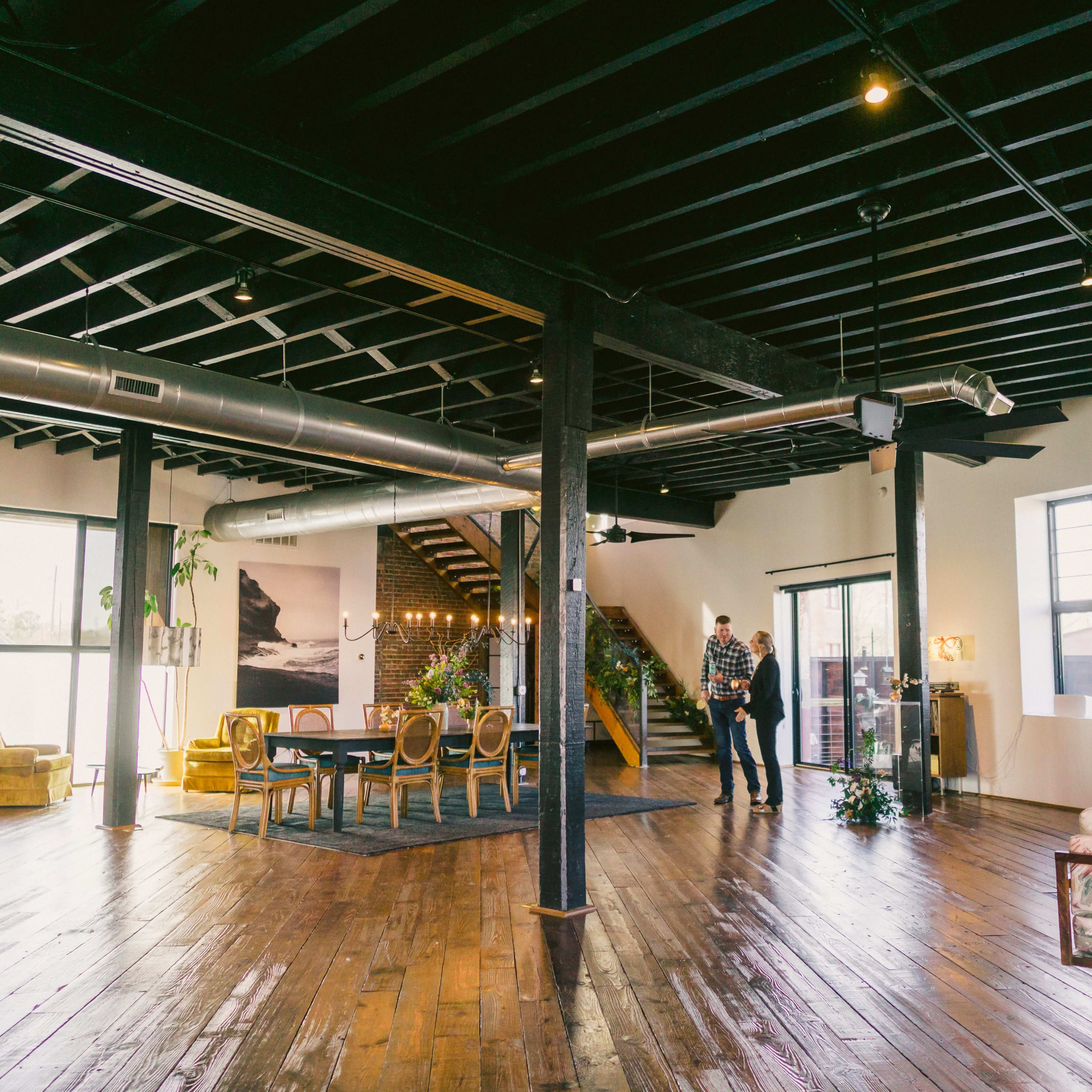 A spacious modern interior with wooden floors, large windows, a dining area, and a staircase leading to an upper level.