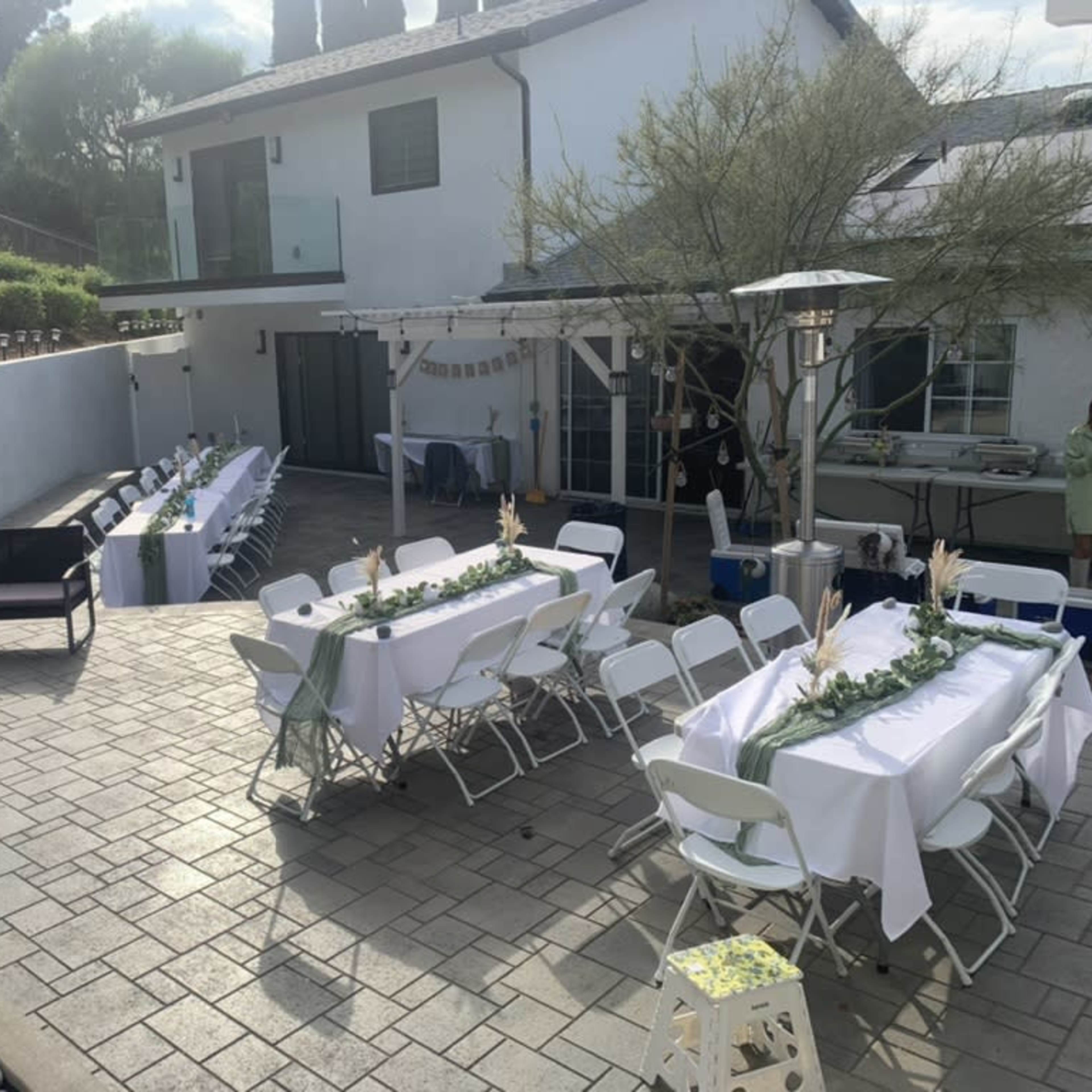 An outdoor dining setup with several tables covered in white tablecloths, arranged alongside a patio surrounded by greenery and residential buildings.