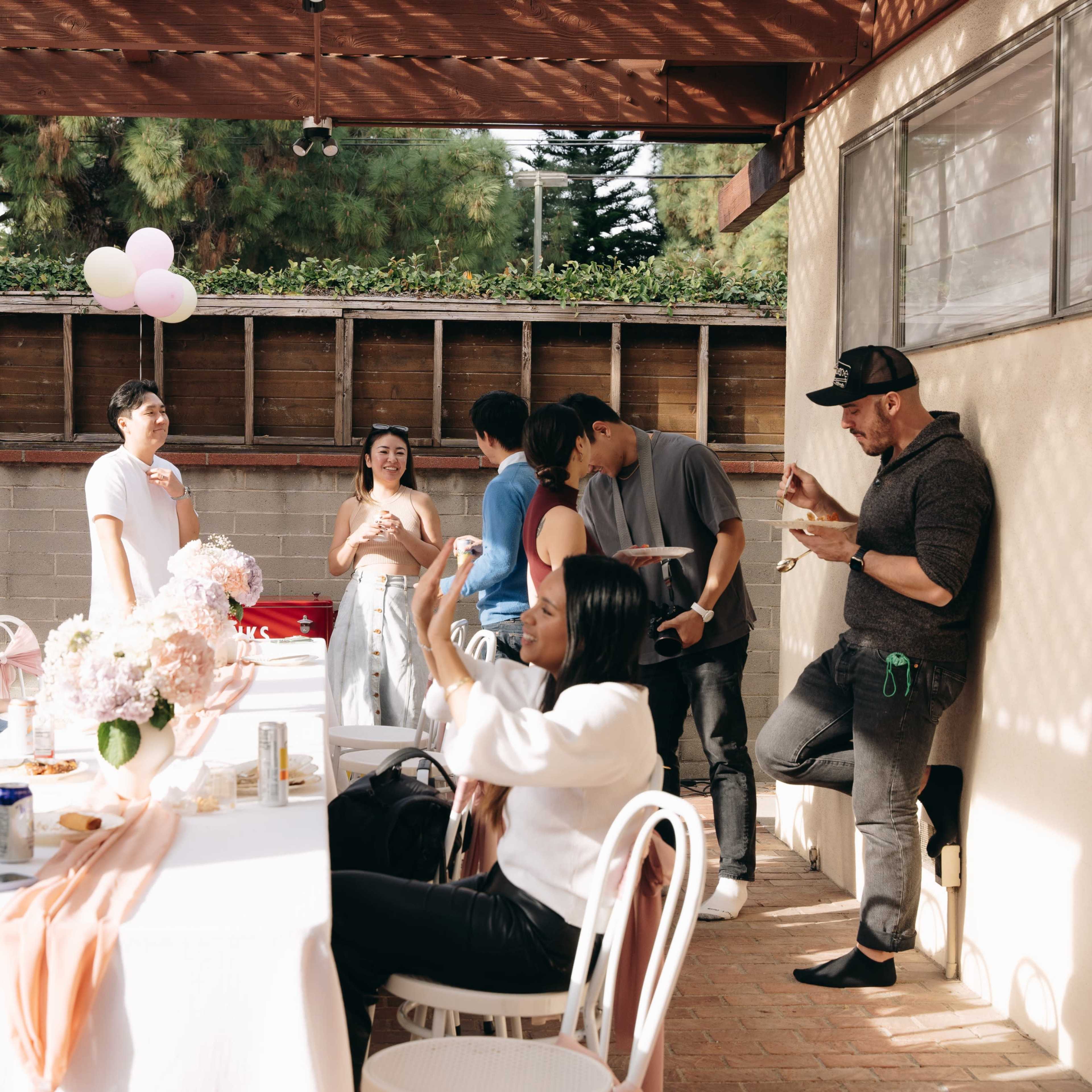 A group of people gather around a table decorated with flowers at a social gathering in a backyard.