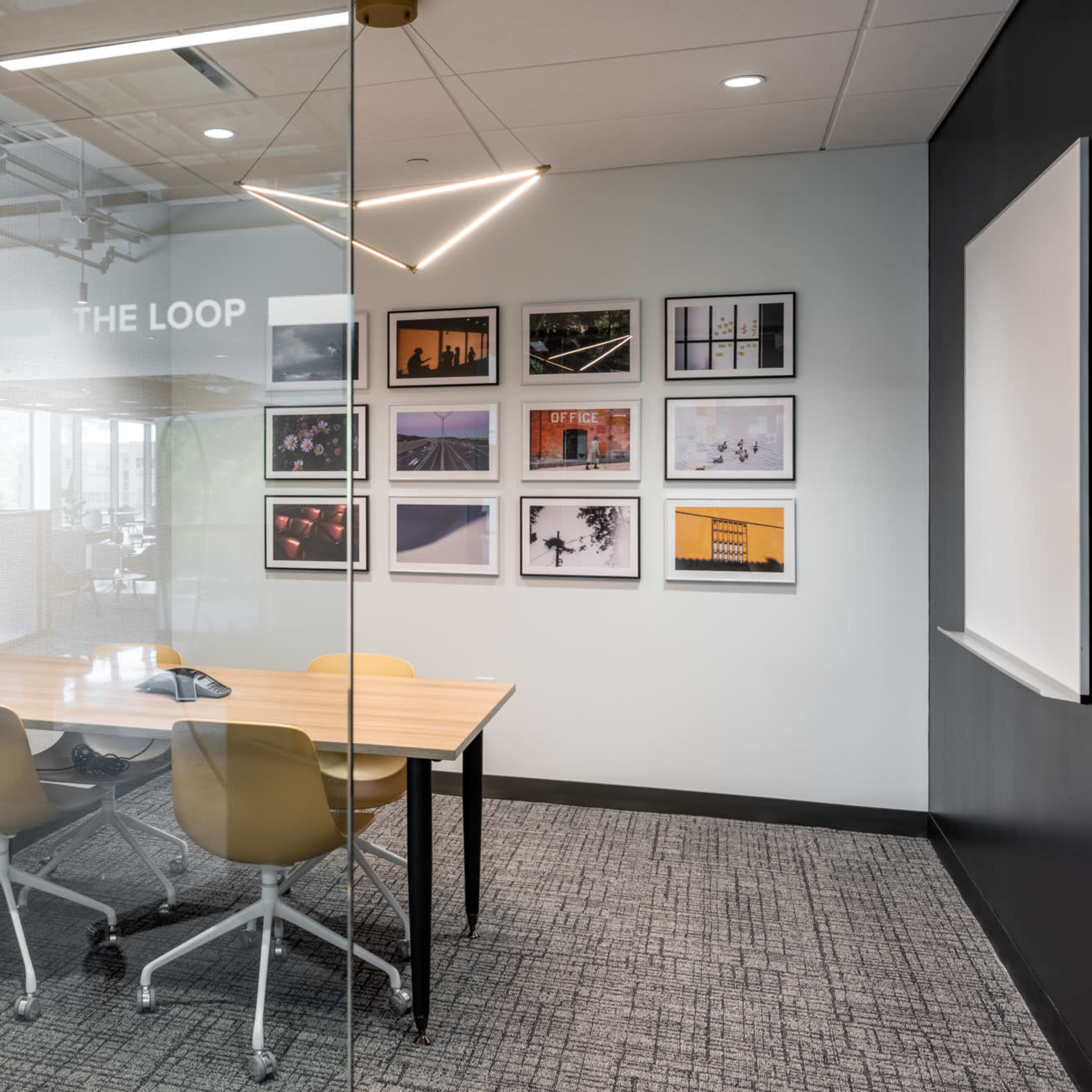 A glass-walled conference room features a wooden table with four chairs and framed photographs on the wall.