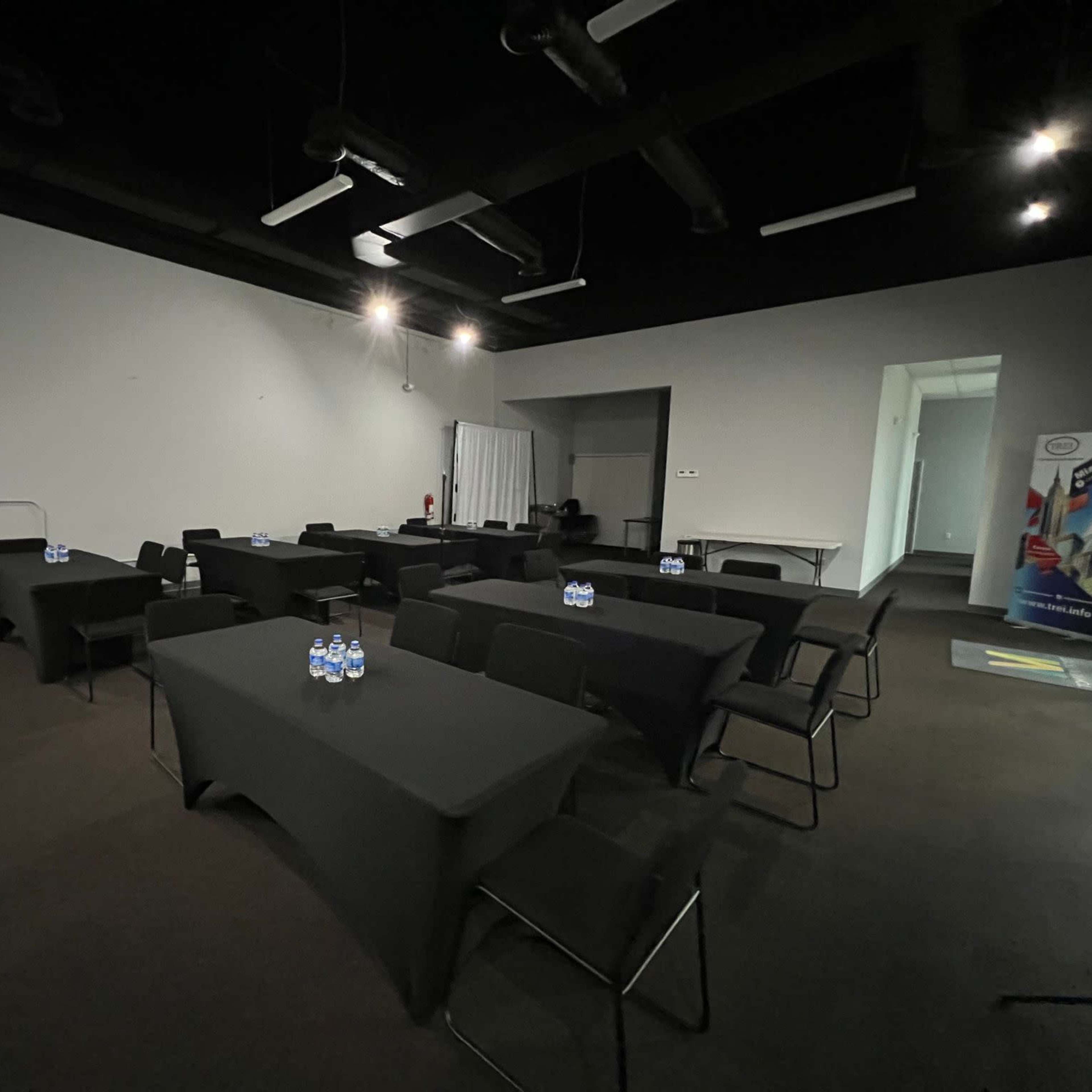 A meeting room with several tables arranged in rows, each with water bottles on top, and minimal lighting.