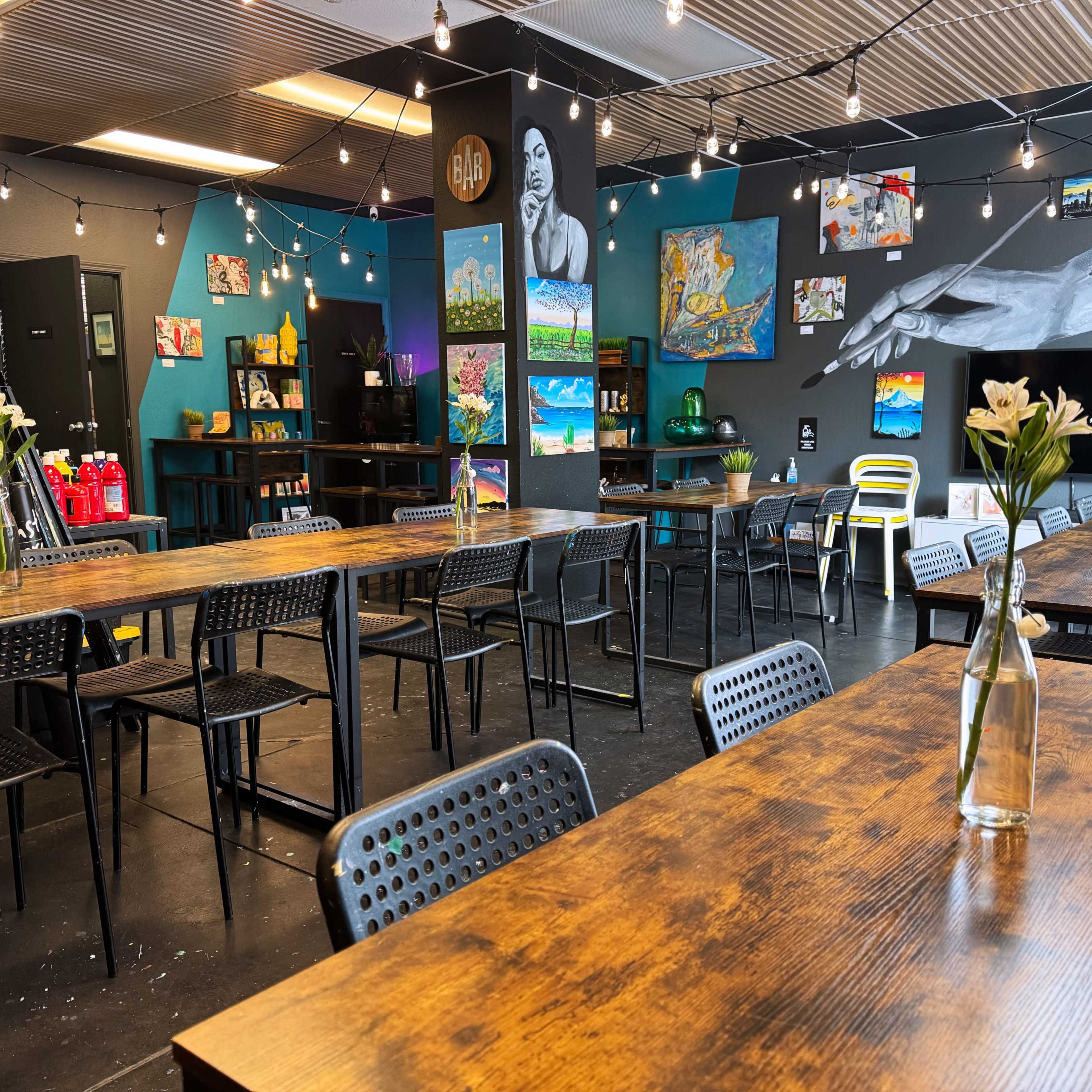 A vibrant dining space with wooden tables, black chairs, and colorful artwork on the walls, illuminated by string lights.