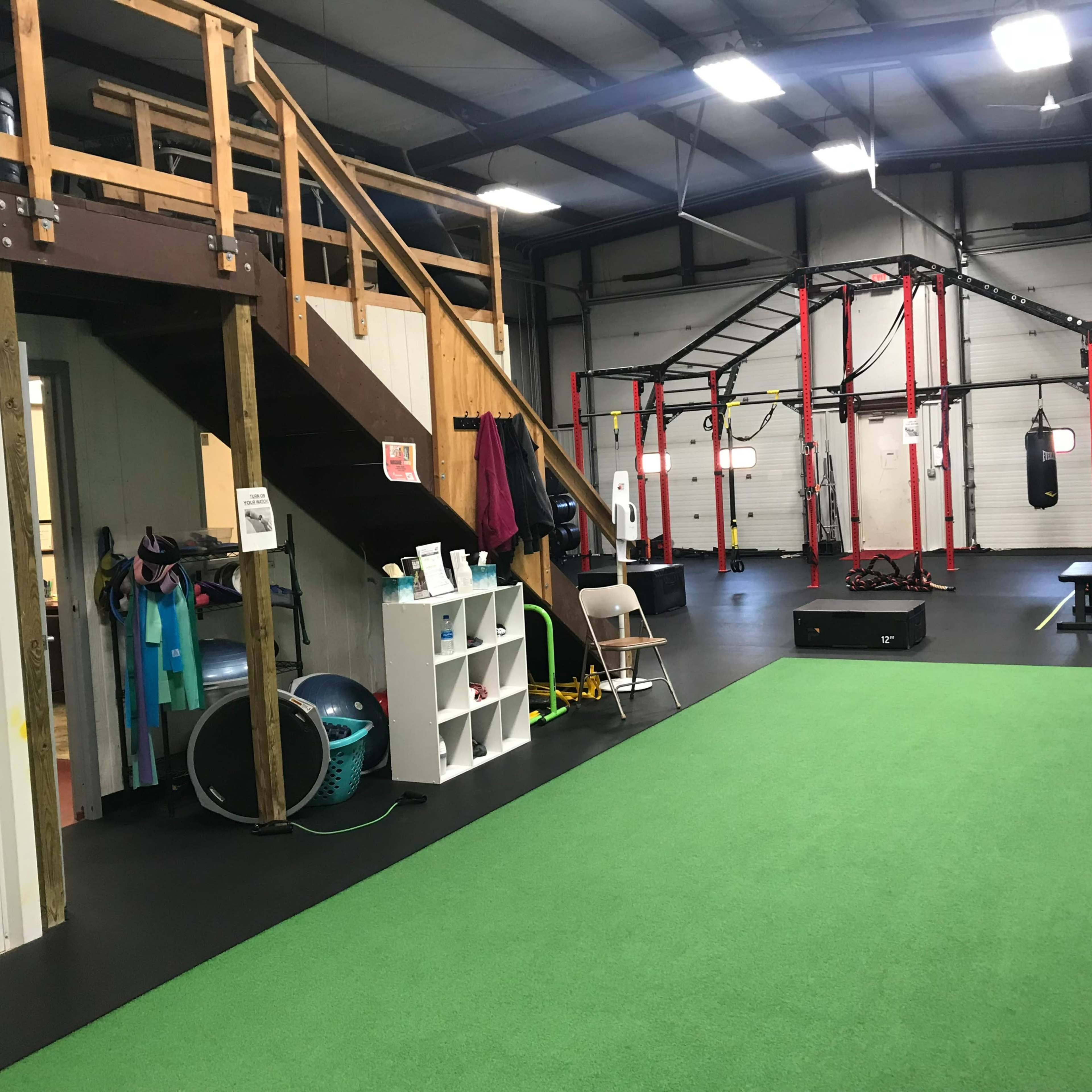 An indoor gym area featuring a turf floor, exercise equipment, a staircase leading to a loft, and various fitness apparatuses suspended from a frame.
