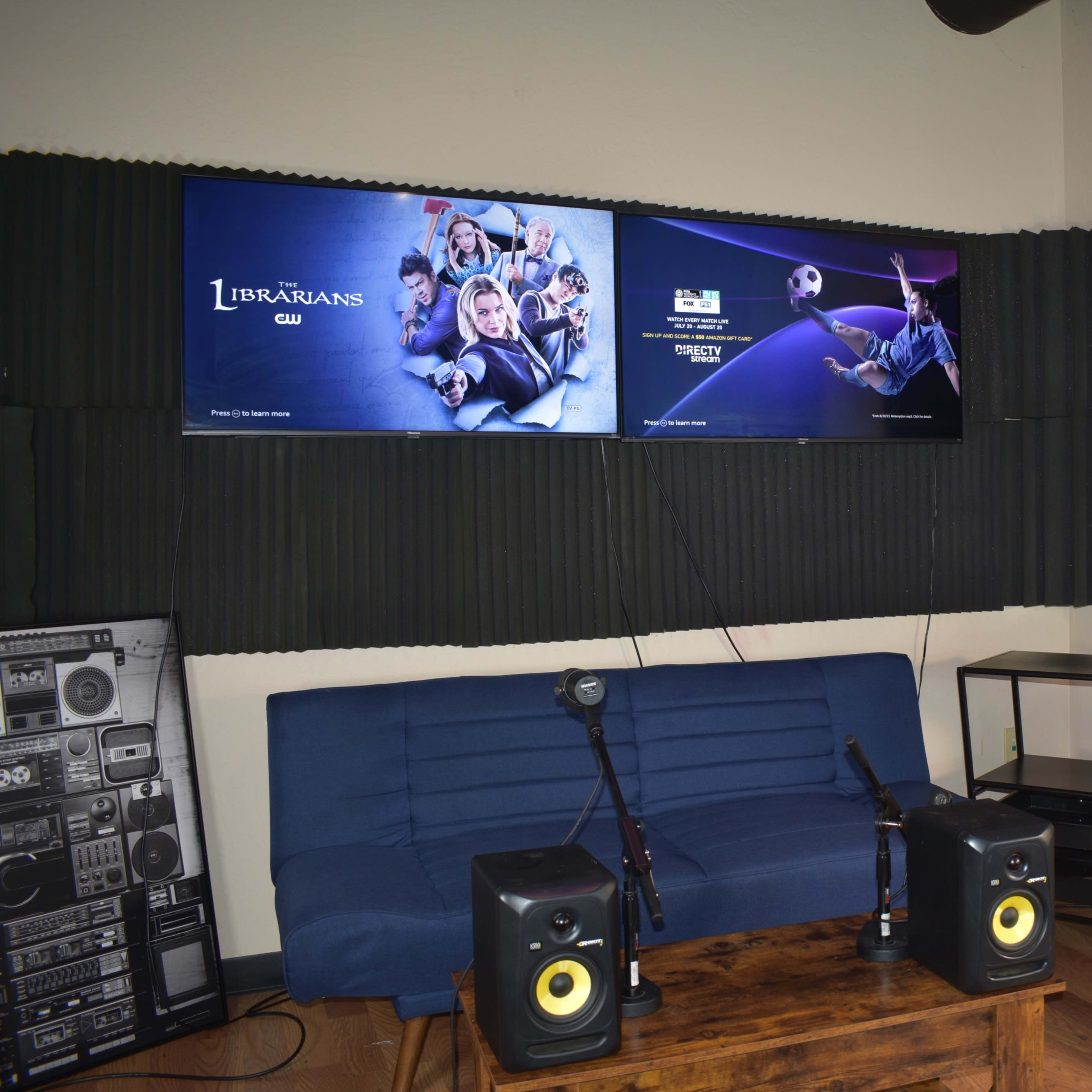 A small studio setup featuring two television screens mounted on a soundproofed wall, a blue sofa, a microphone on a stand, and audio equipment.