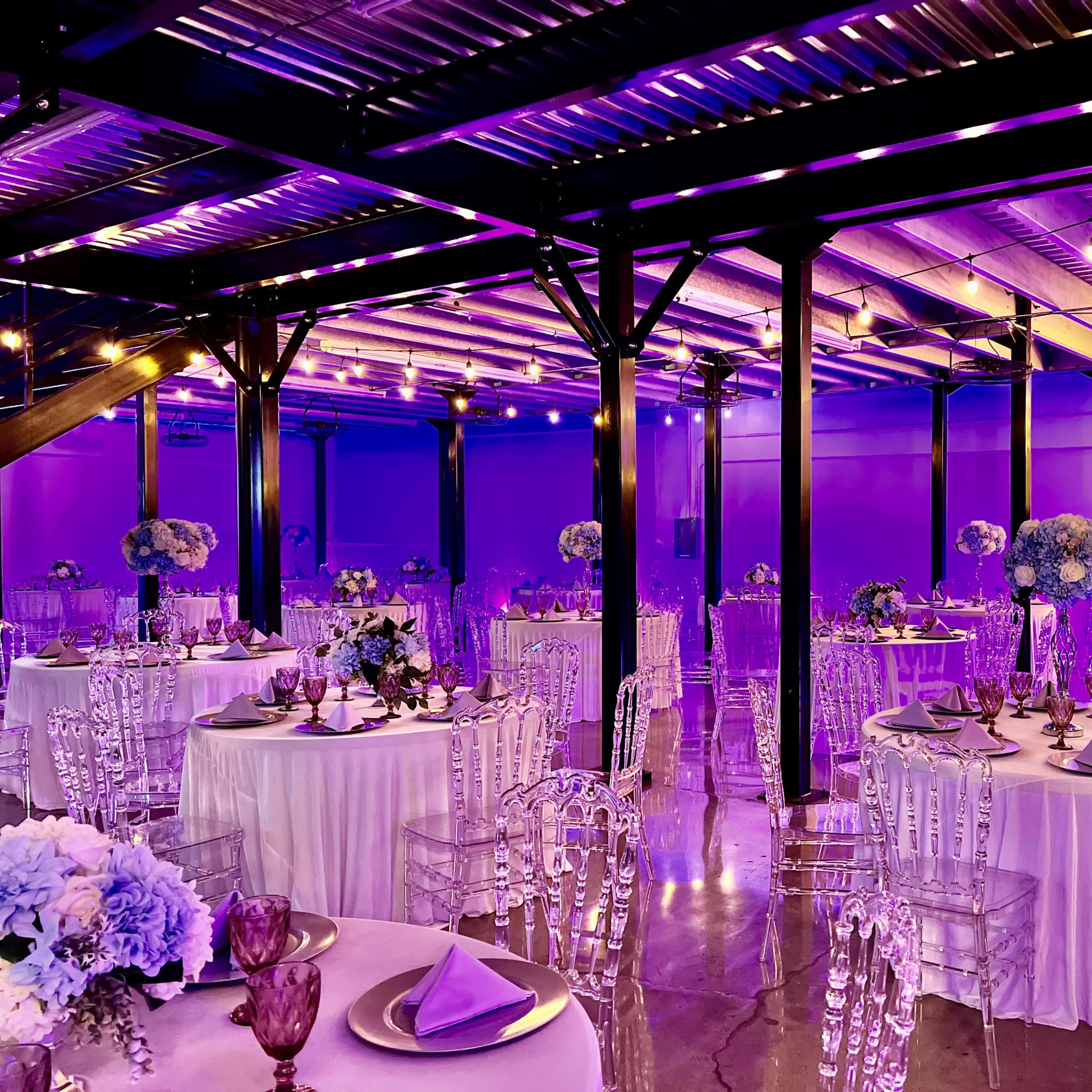 A modern event space features round tables with white tablecloths, clear acrylic chairs, and purple lighting, along with a bar area illuminated by bright letters.