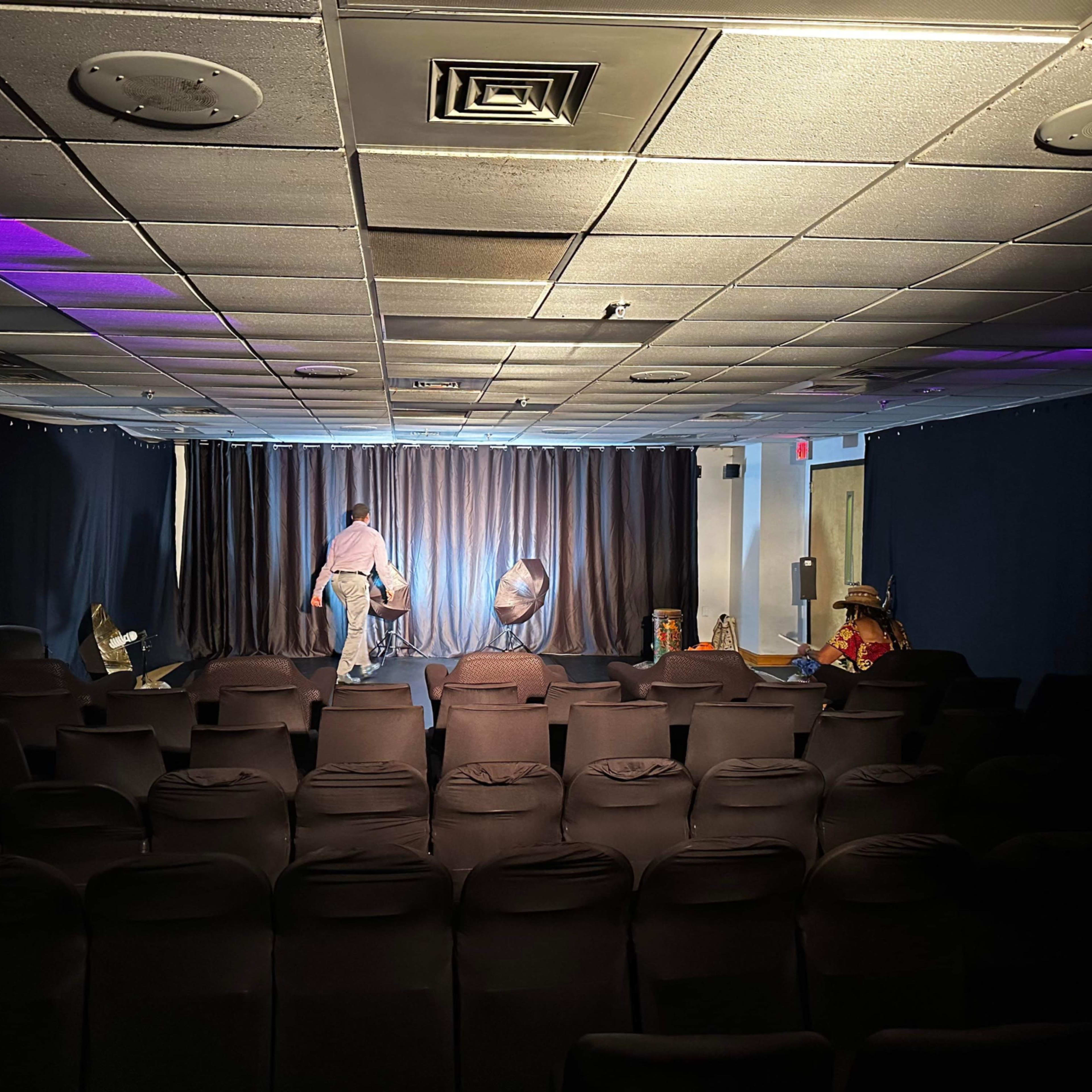 A small theater space with black chairs, a stage draped in curtains, and a performer setting up props under dim lighting.