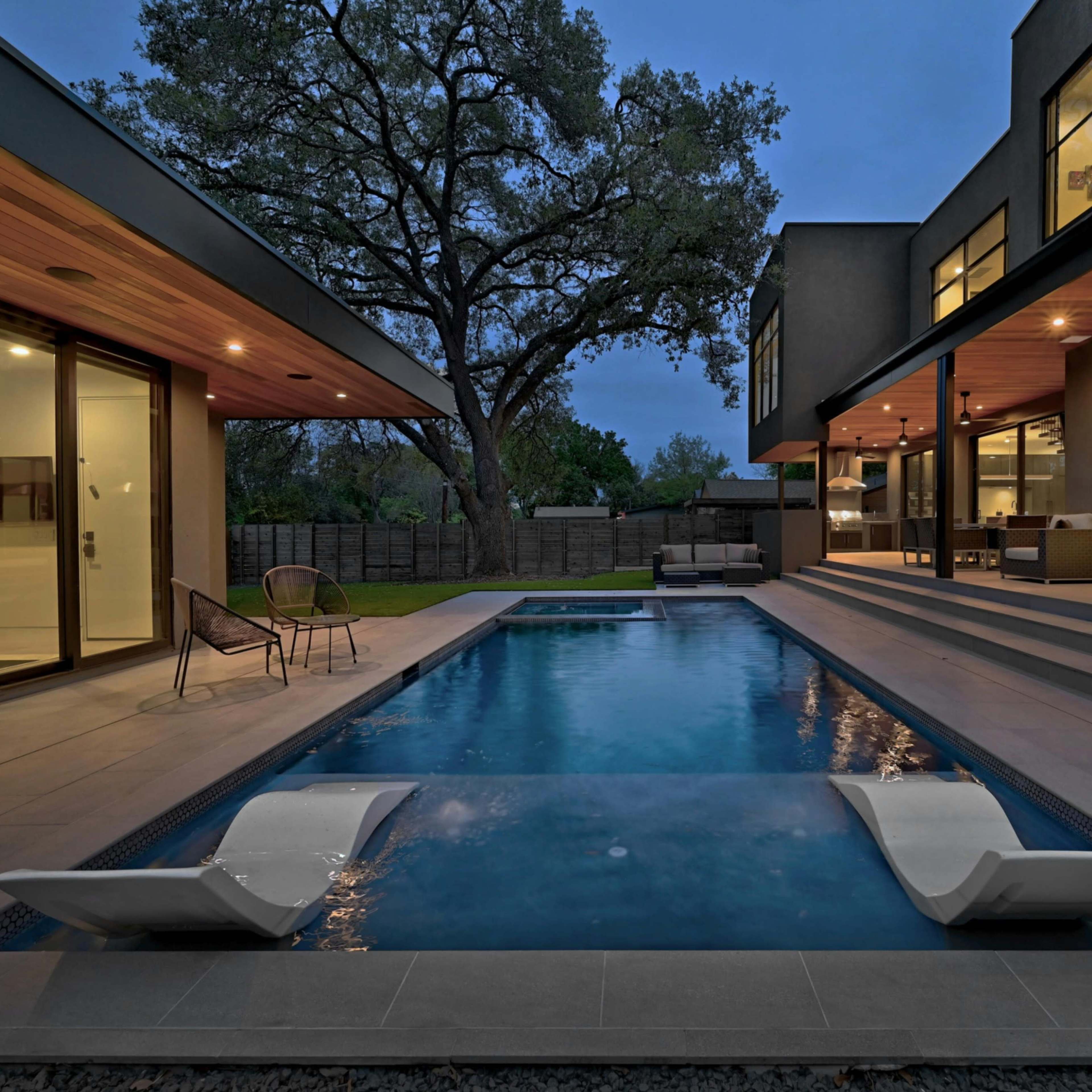 A modern home features a sleek pool with lounge chairs next to a spacious patio and large glass doors leading indoors at twilight.
