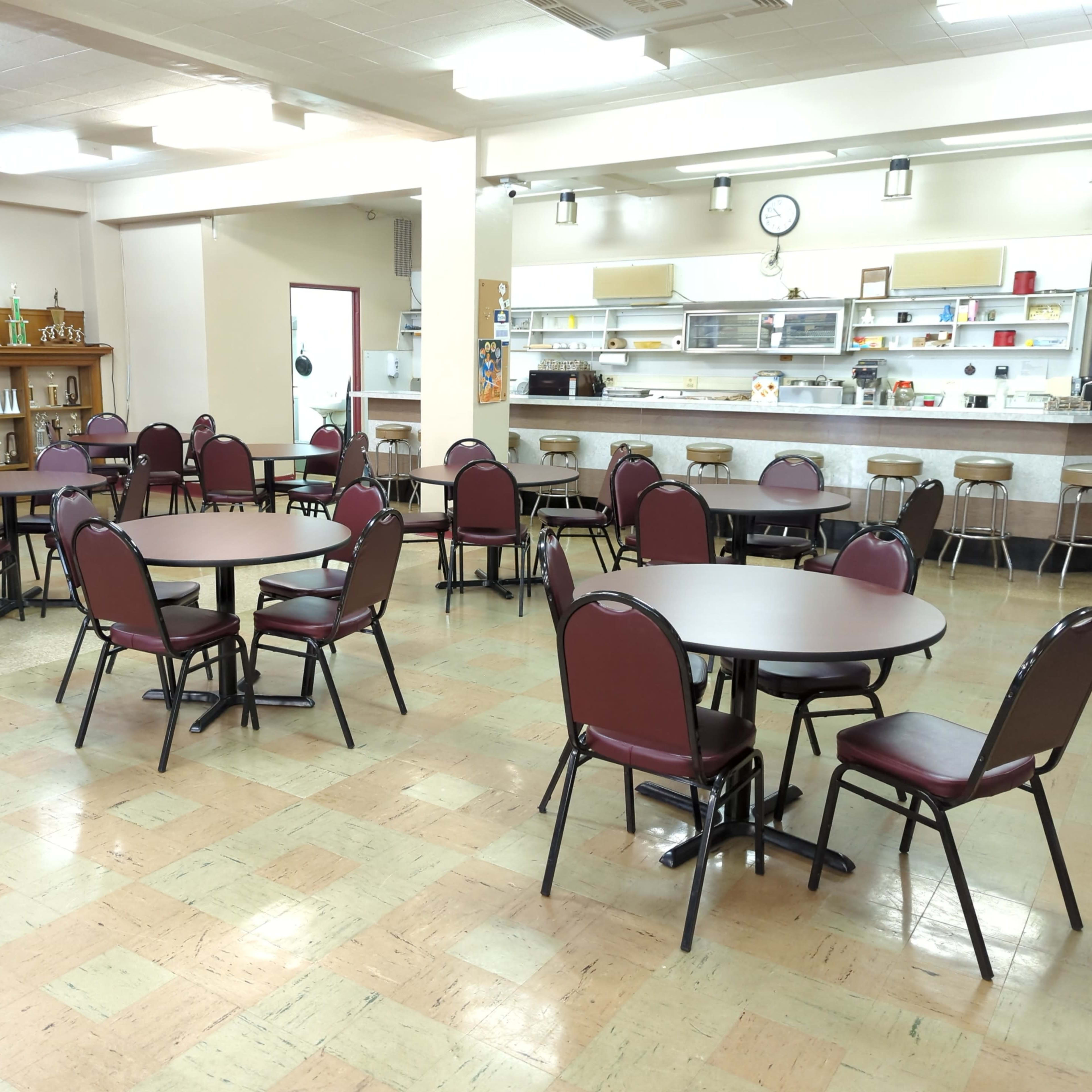 A dining area with several round tables and chairs arranged in a room featuring a counter and kitchen space in the background.