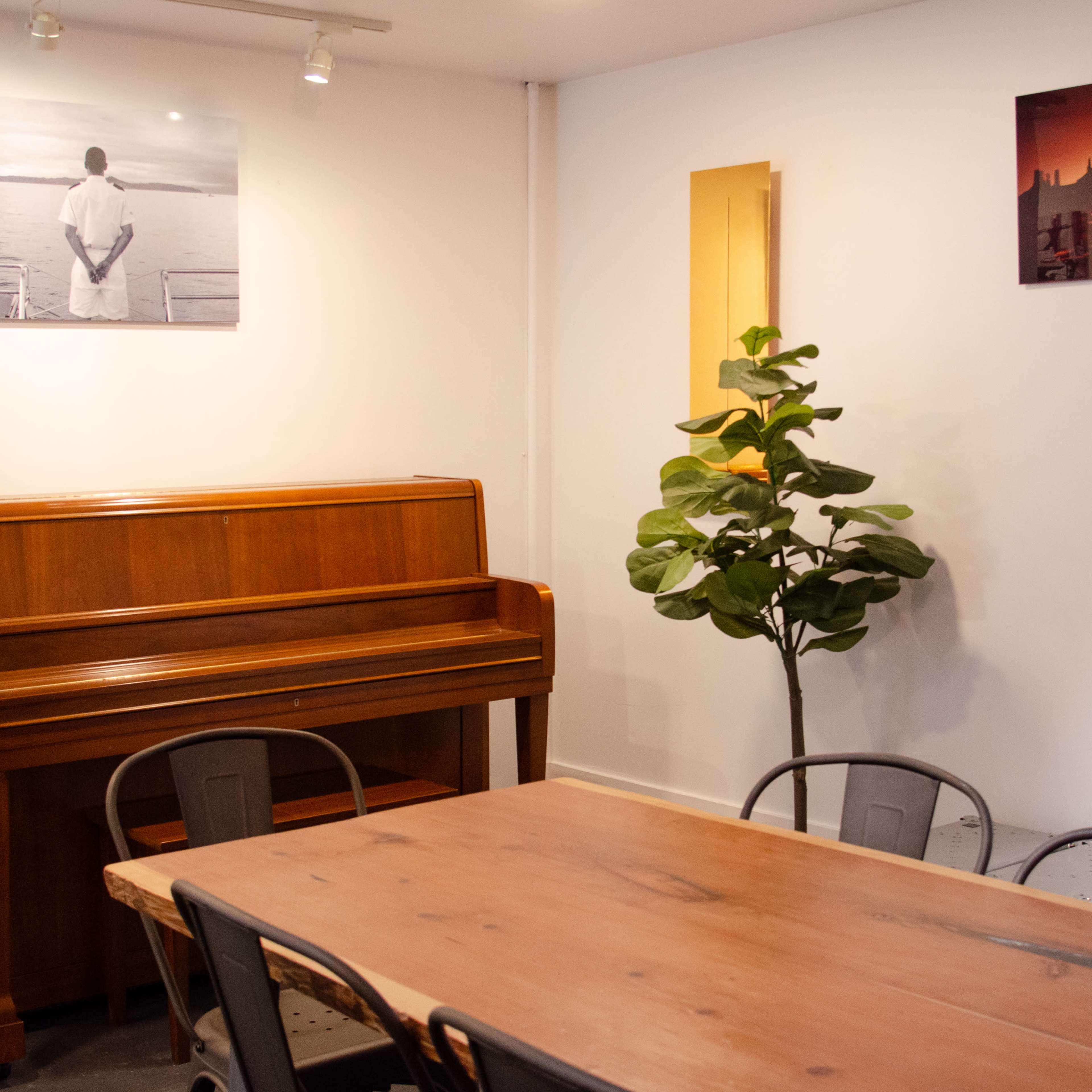 A room with a wooden piano, a large wooden table with metal chairs, and two framed photographs on the wall above the piano.