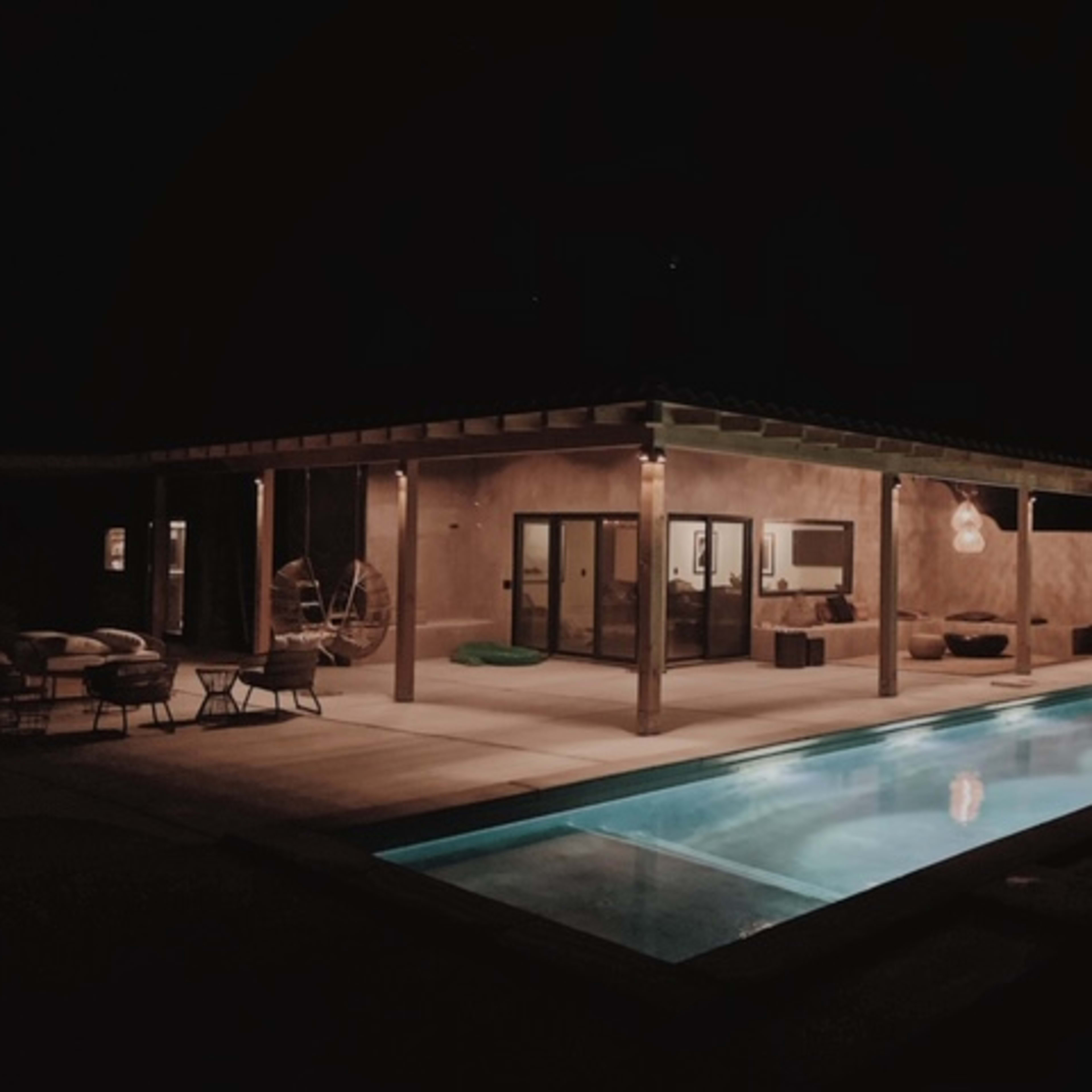 A modern outdoor space featuring a pool and a covered patio with furniture, illuminated softly at night.