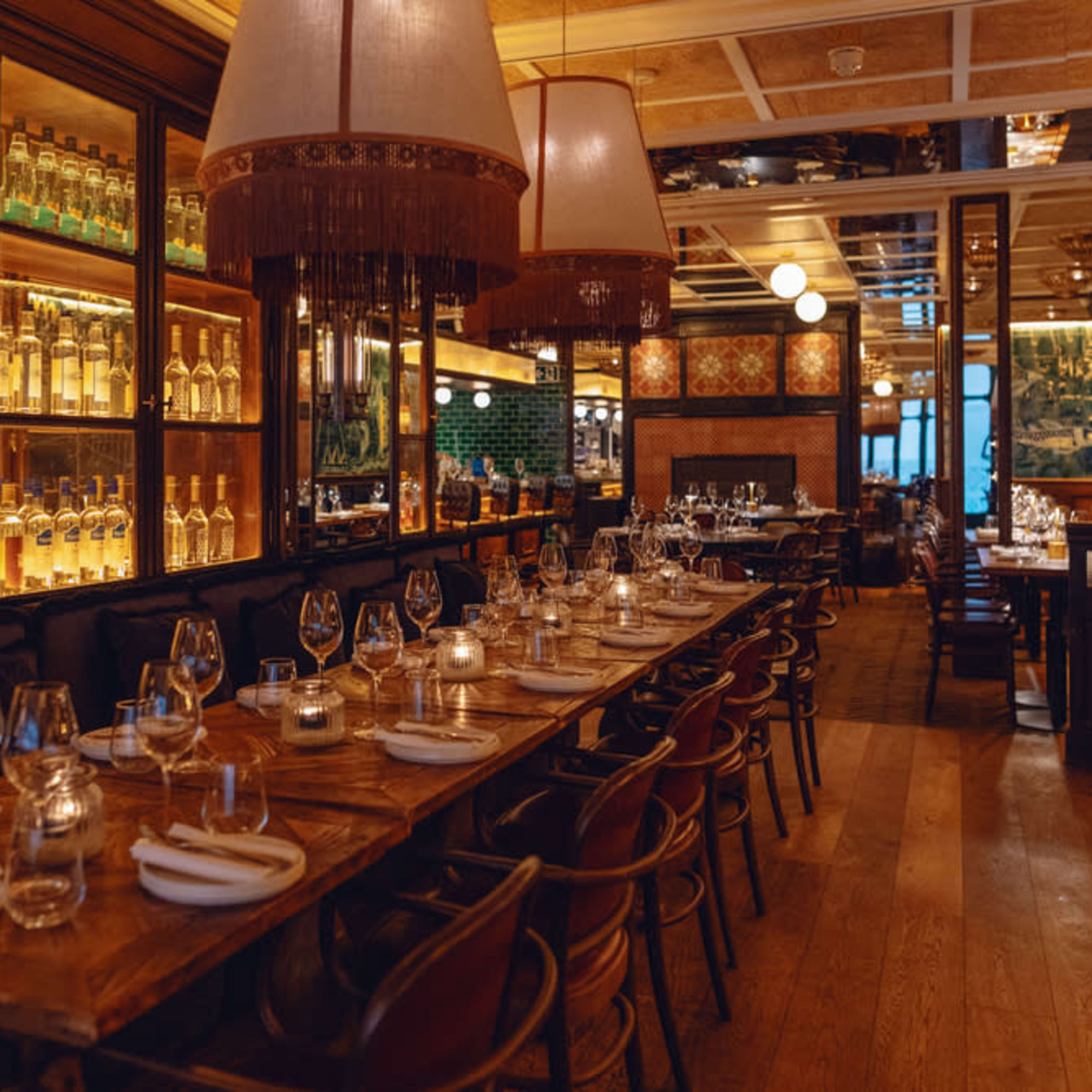A warmly lit restaurant interior with a long wooden table set for dining, surrounded by shelves displaying bottles and stylish pendant lights.
