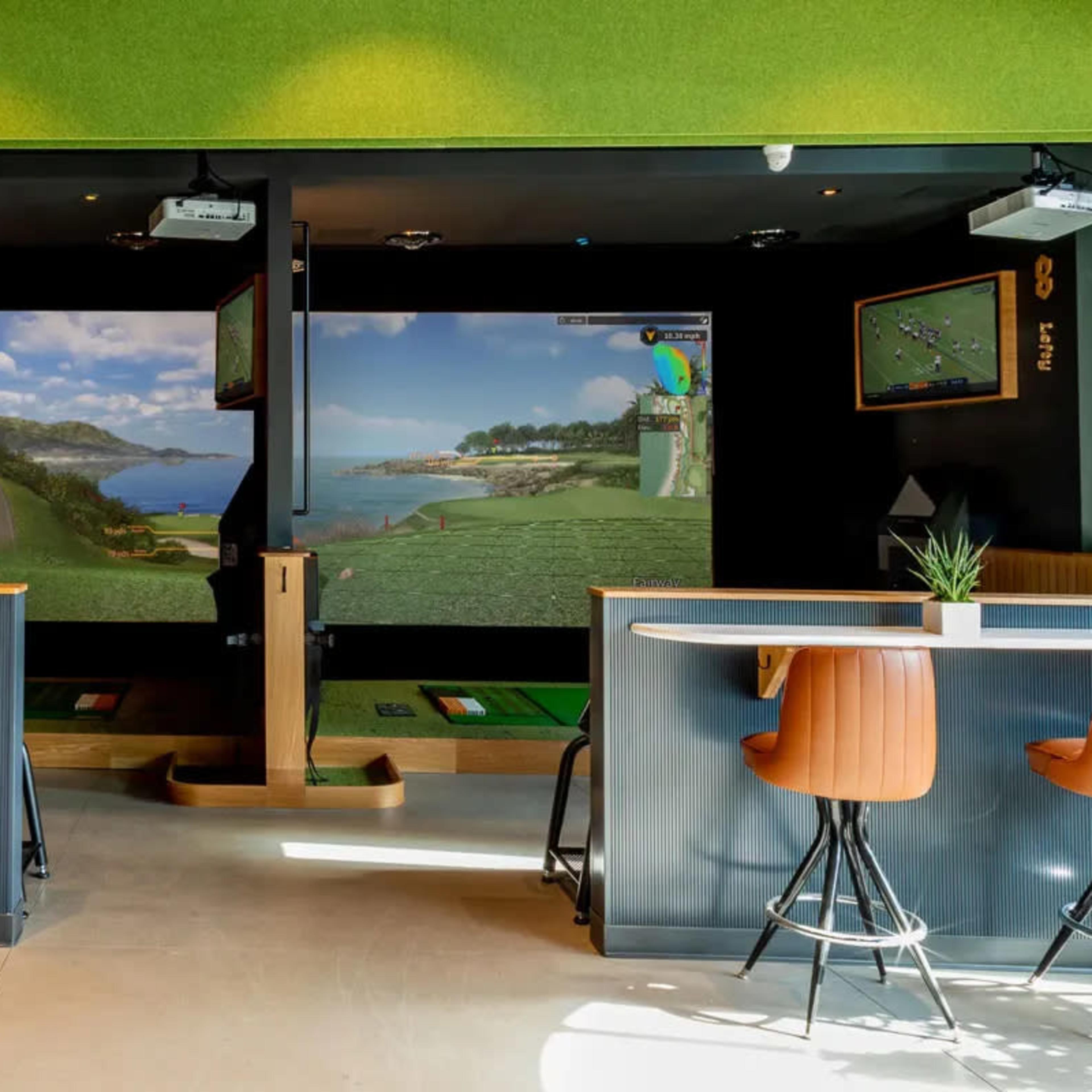 An indoor golf simulator space with two large screens displaying golf course scenes, along with golf mats and seating around a bar area.