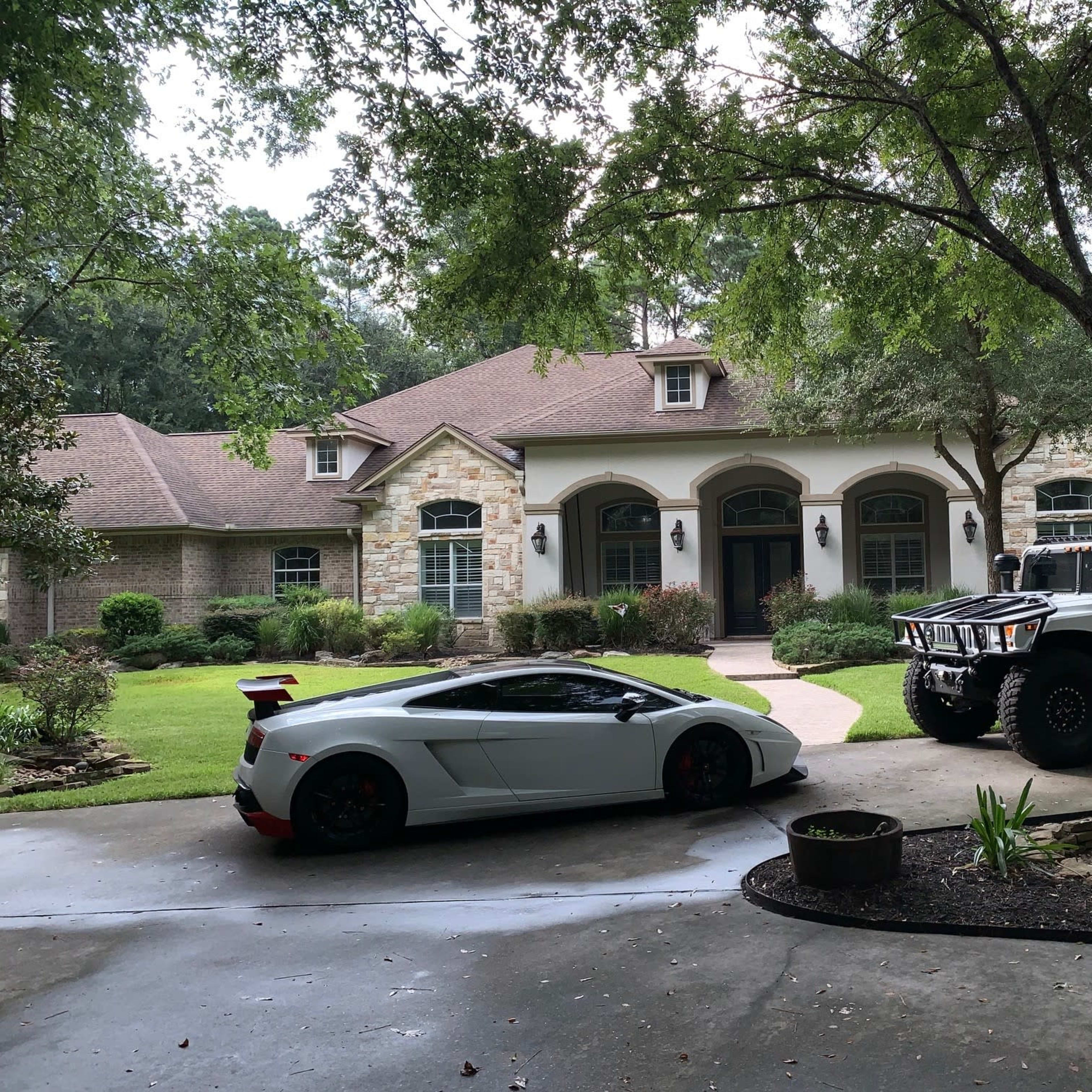 A white Lamborghini and a white Hummer are parked in front of a large, stone-accented residential house surrounded by greenery.