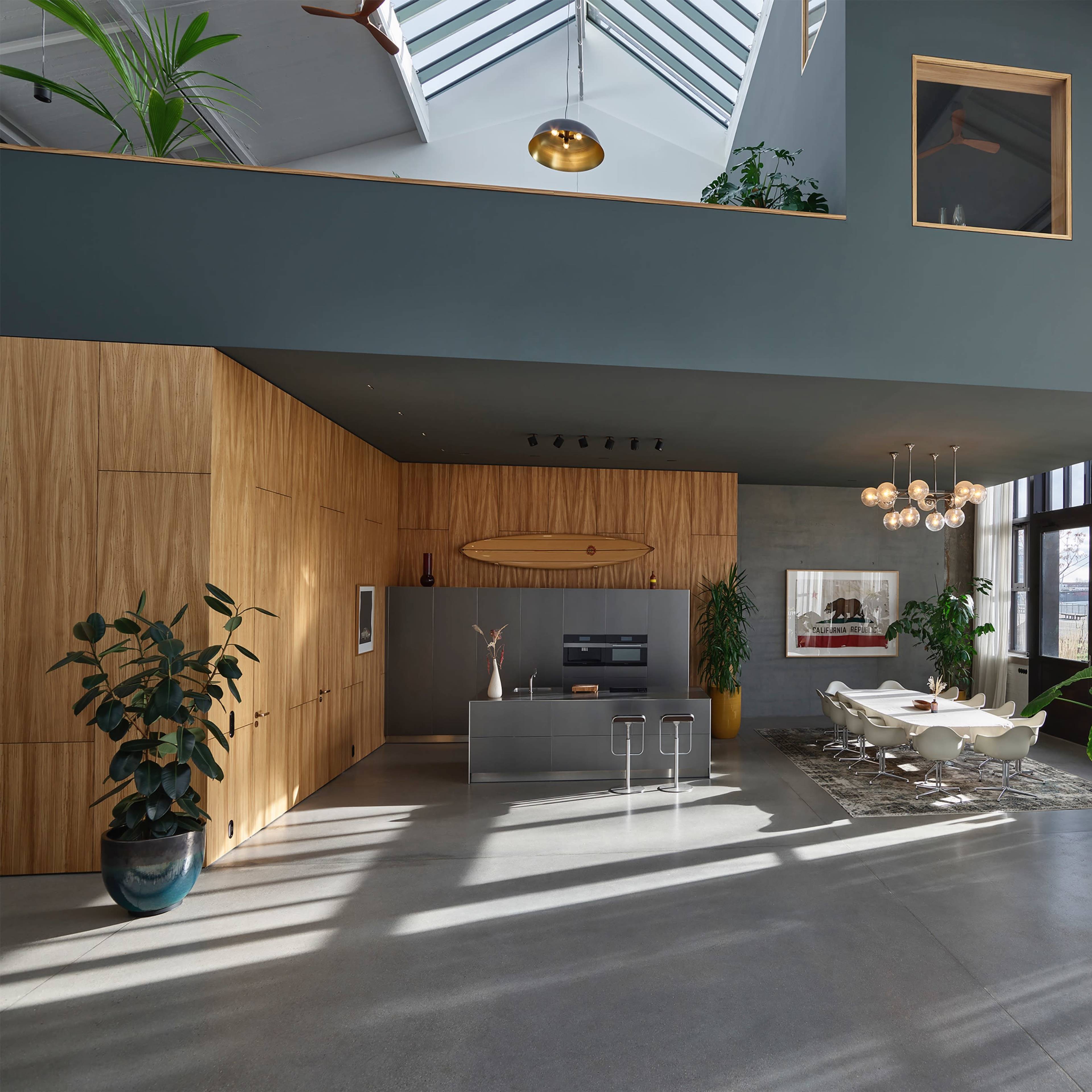 A modern, spacious living area featuring wooden cabinetry, large windows, and contemporary furnishings, with plants positioned throughout the space.
