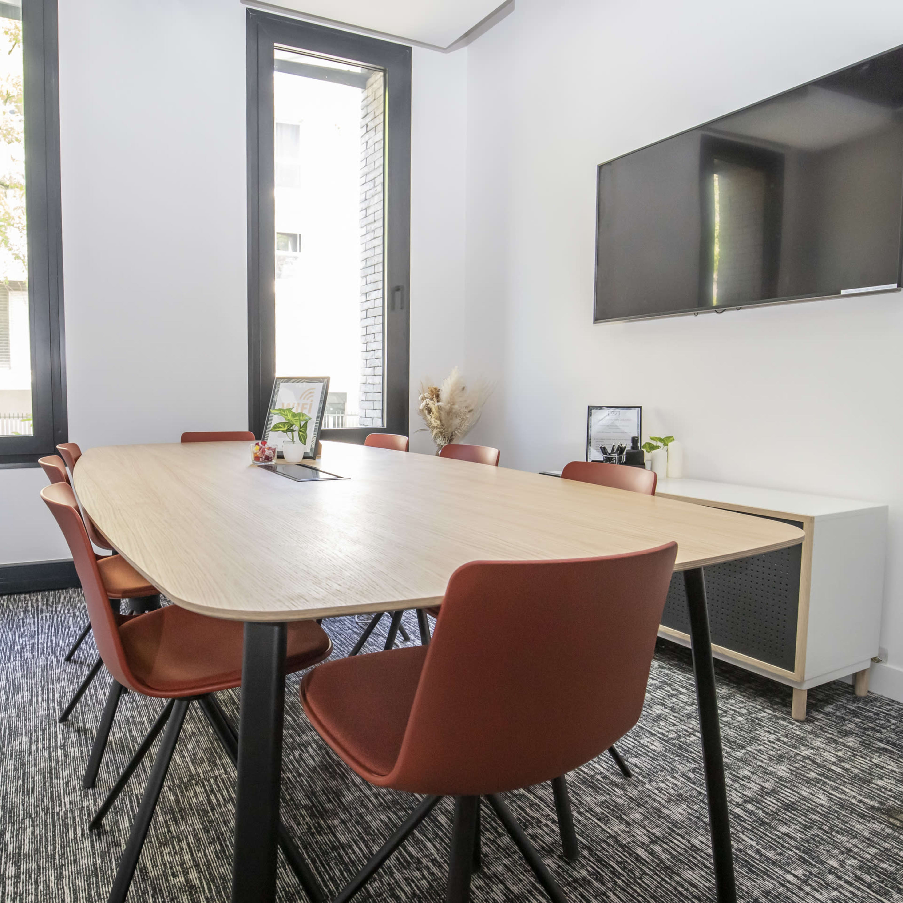 A modern meeting room features a large wooden table surrounded by eight chairs, with large windows allowing natural light to illuminate the space.