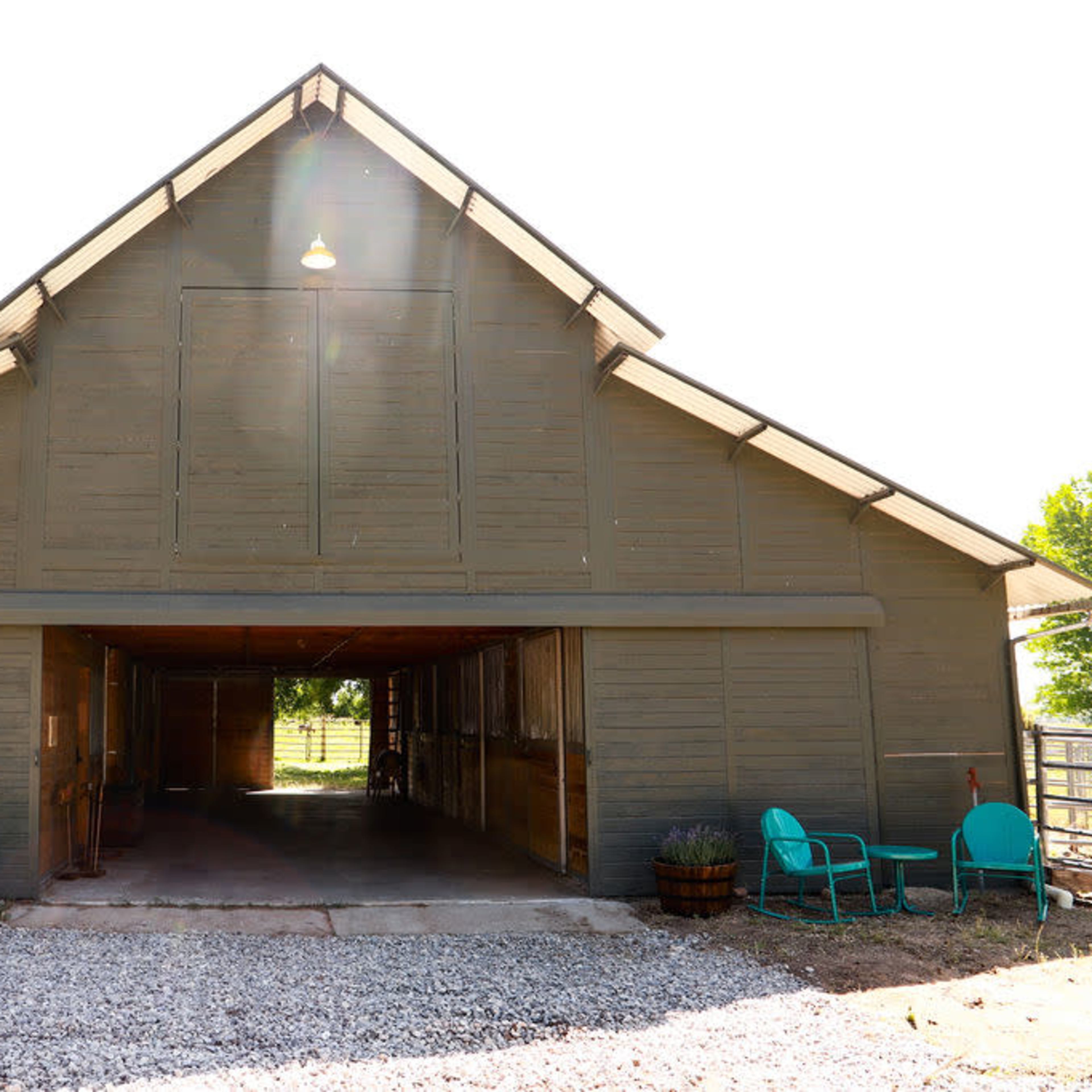 A large, wooden barn with an open front and a gravel yard features two blue chairs on either side.