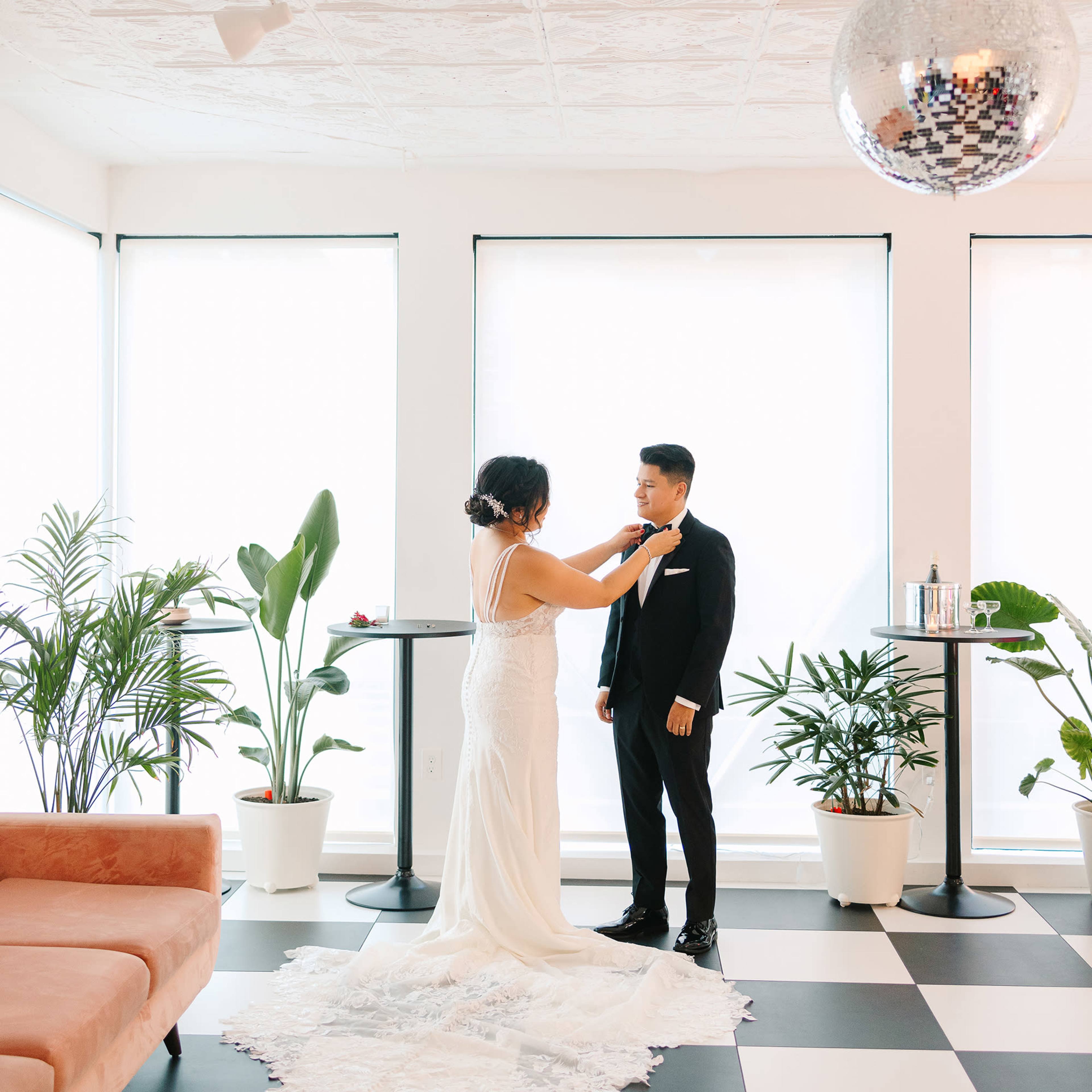 A bride adjusts the tie of a groom in a bright room with large windows and a disco ball, surrounded by indoor plants and a pink sofa.