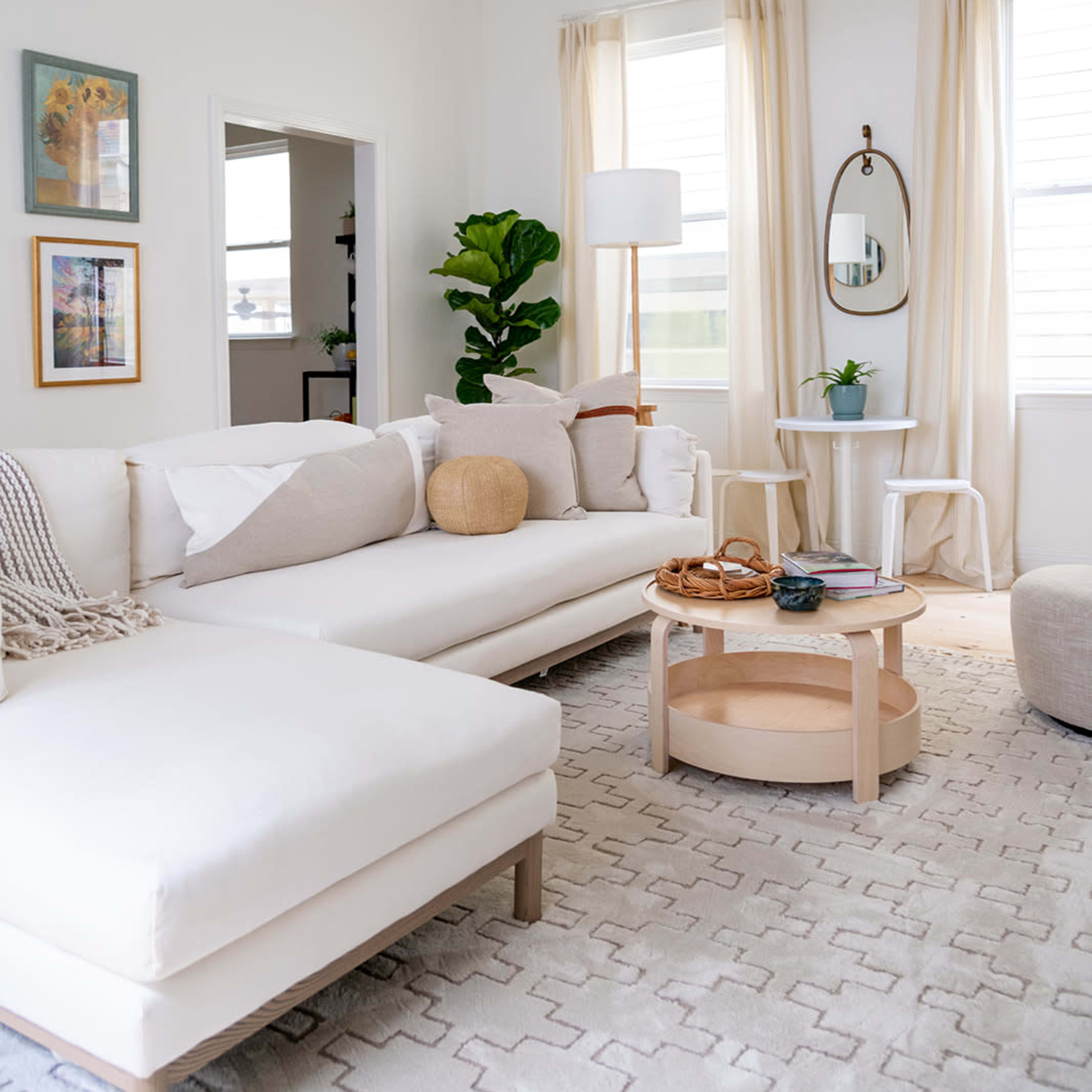 A bright living room features a white sectional sofa, a round coffee table, and a floor lamp, with large windows allowing natural light to illuminate the space.