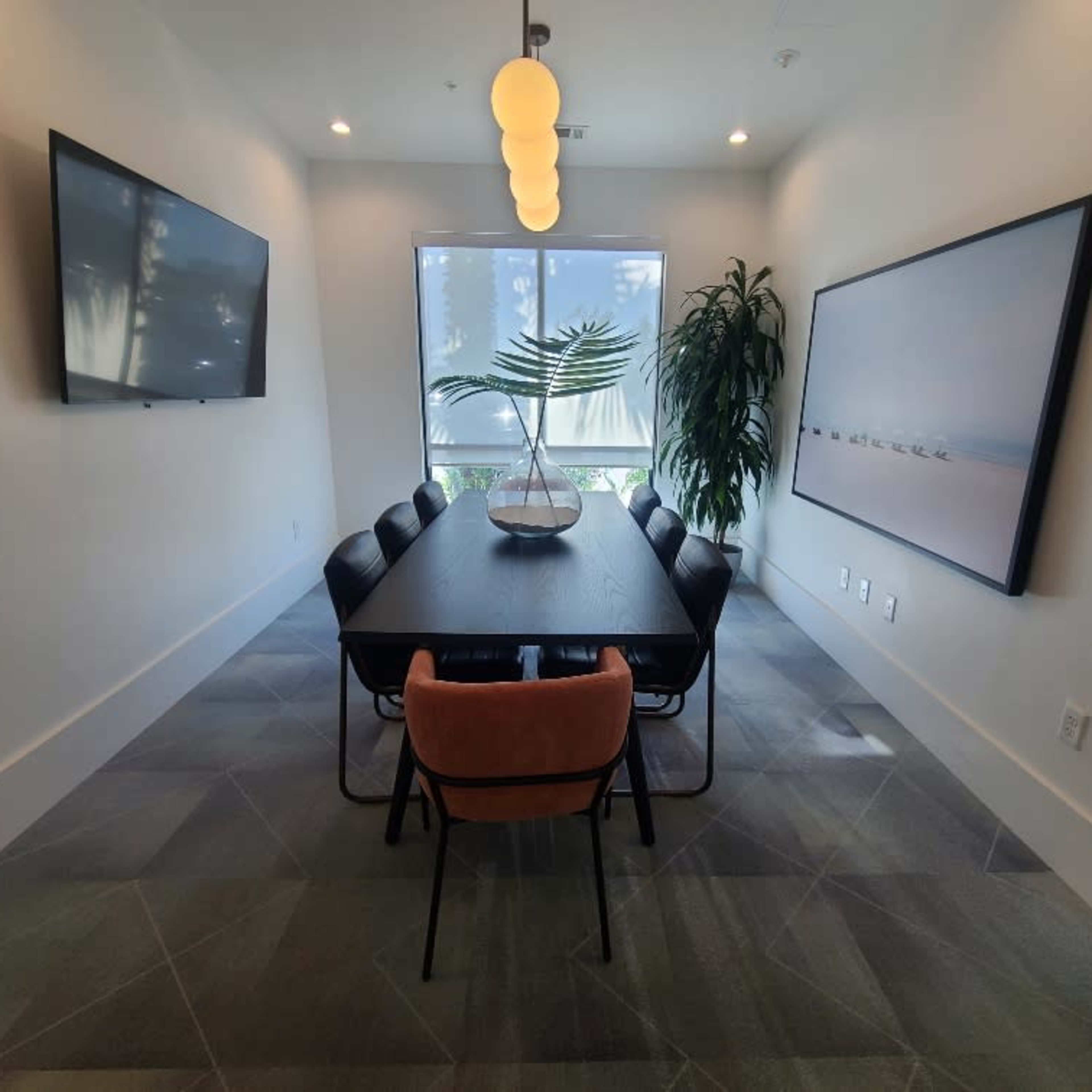 A modern conference room features a long table surrounded by chairs, with a TV on the wall and a large window letting in natural light.