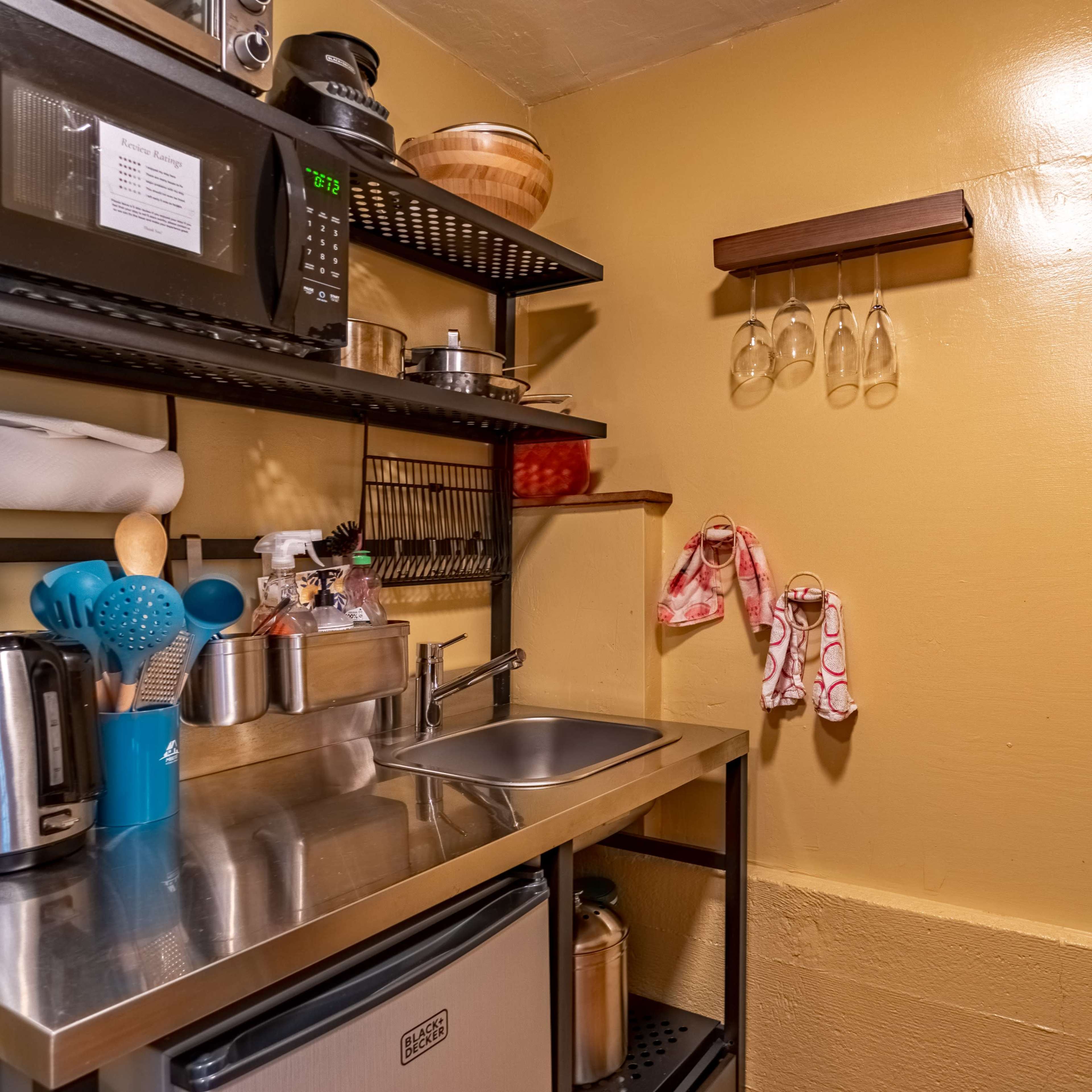 A compact kitchen area featuring stainless steel countertops, a small sink, a microwave, and shelves displaying kitchen utensils and glassware.