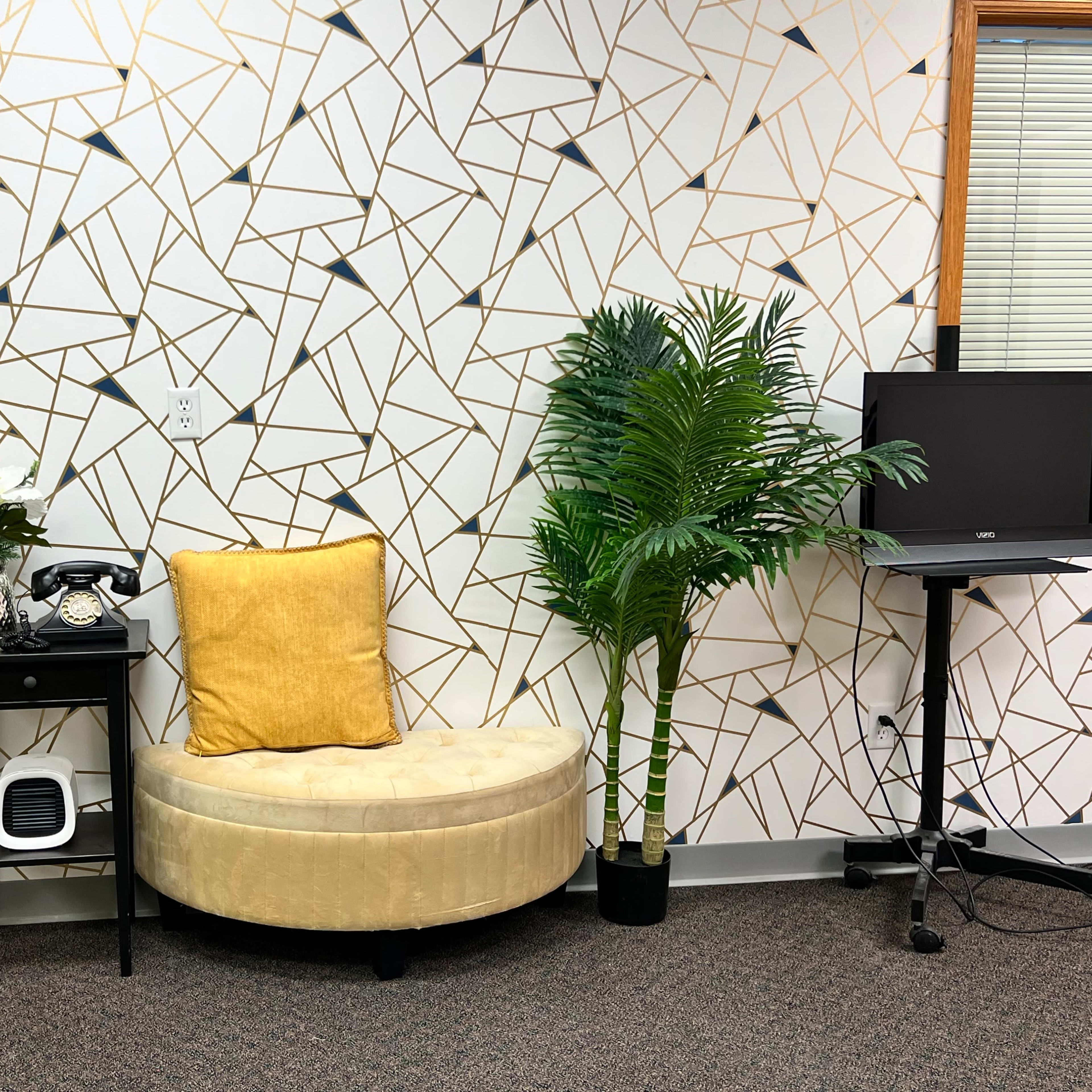 A circular yellow seat is placed next to a small black table with a telephone and a decorative plant, set against a geometric patterned wall and accompanied by a tall artificial palm.