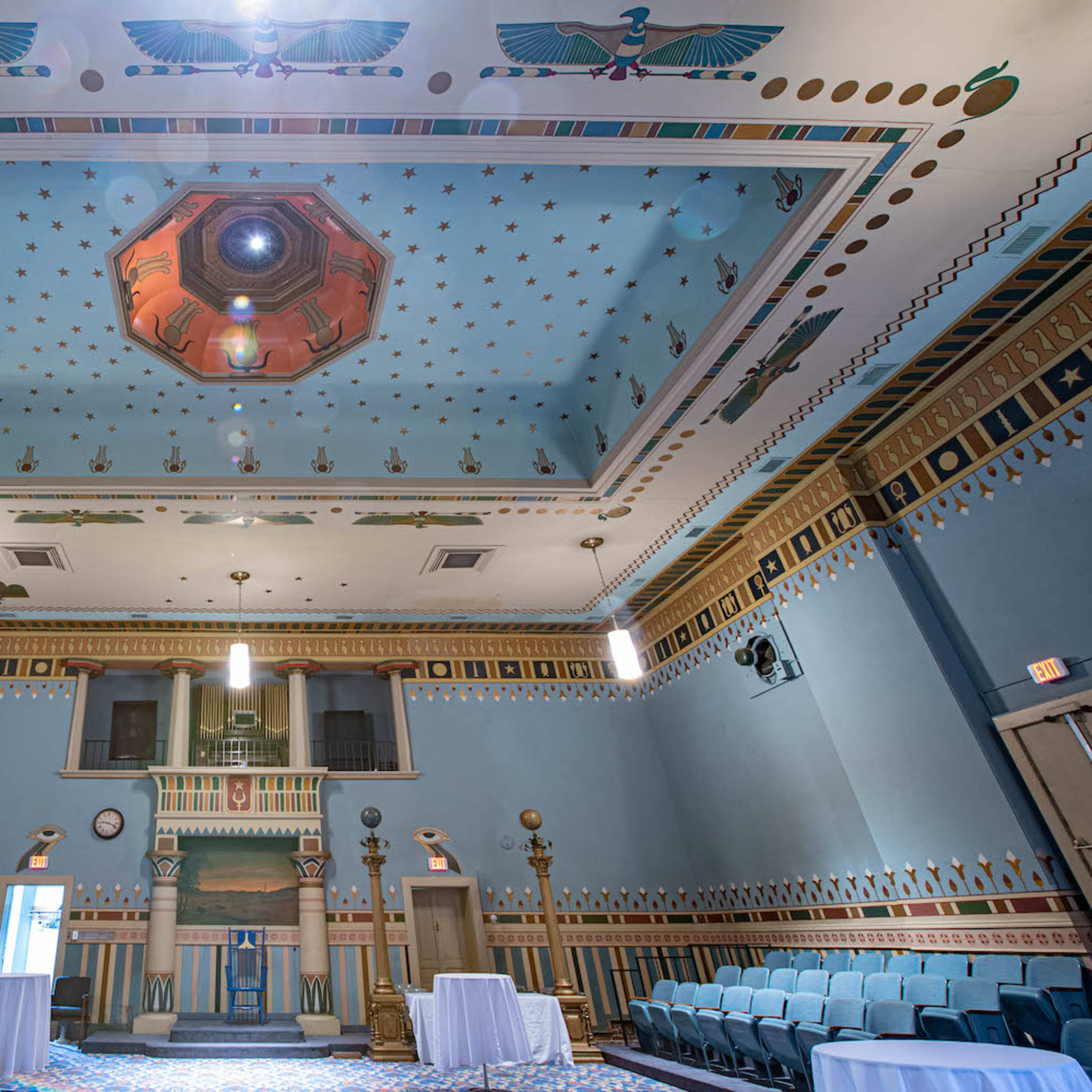 A spacious hall with a decorated ceiling featuring geometric patterns, flanking columns, and tables set up in the center.