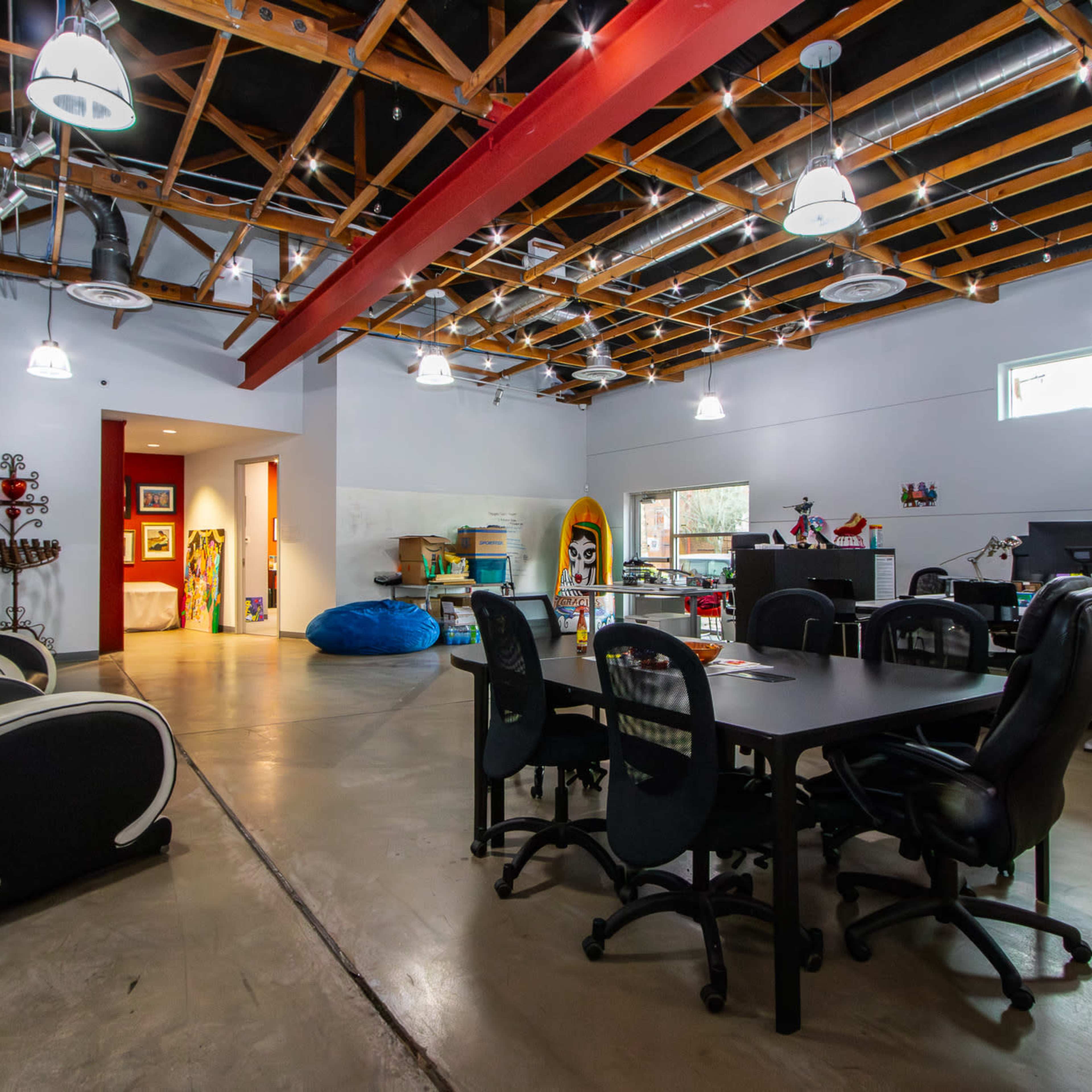 A spacious office area with a concrete floor, large windows, and a wood-beamed ceiling, featuring several desks, chairs, and a blue bean bag.