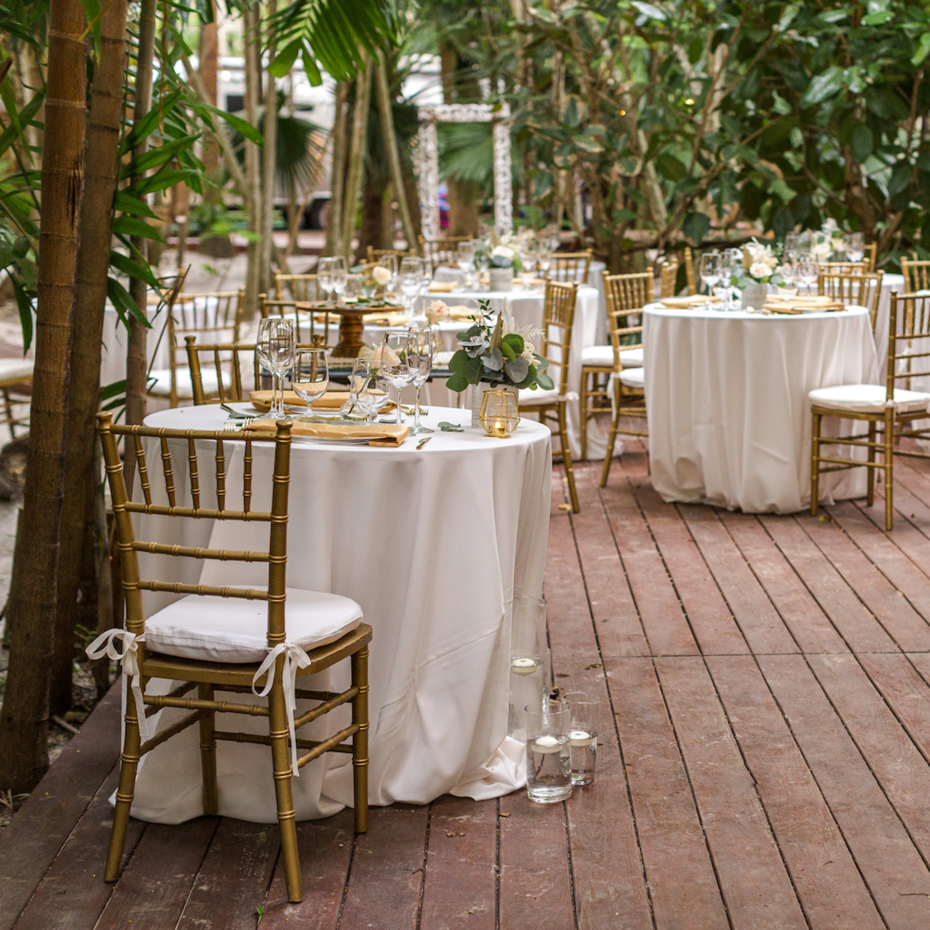 A series of elegantly set dining tables with white tablecloths and gold chairs arranged on a wooden deck surrounded by lush greenery.