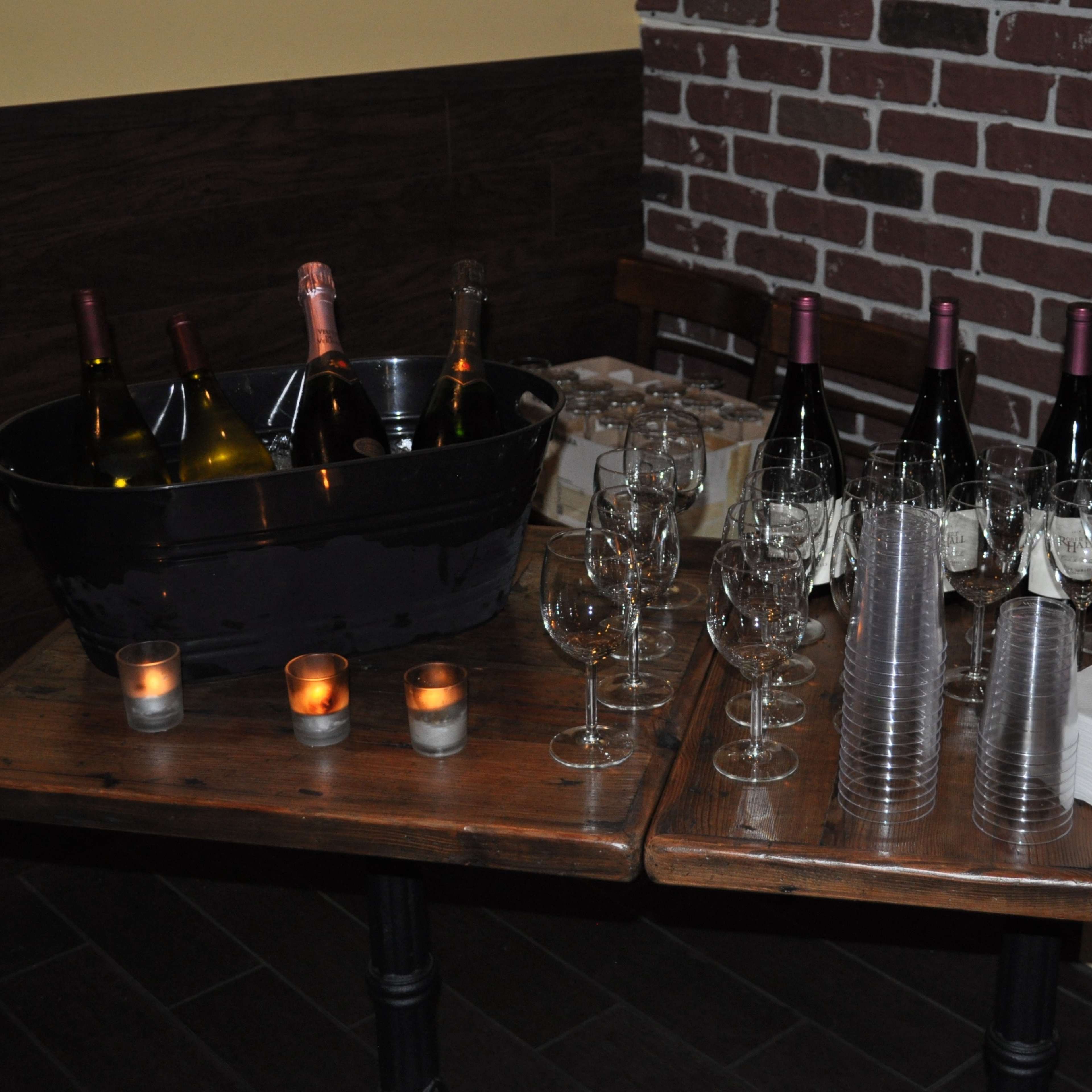 A wooden table displays a bucket of wine bottles, neatly arranged wine glasses, and stacked napkins, with candles providing ambient lighting.