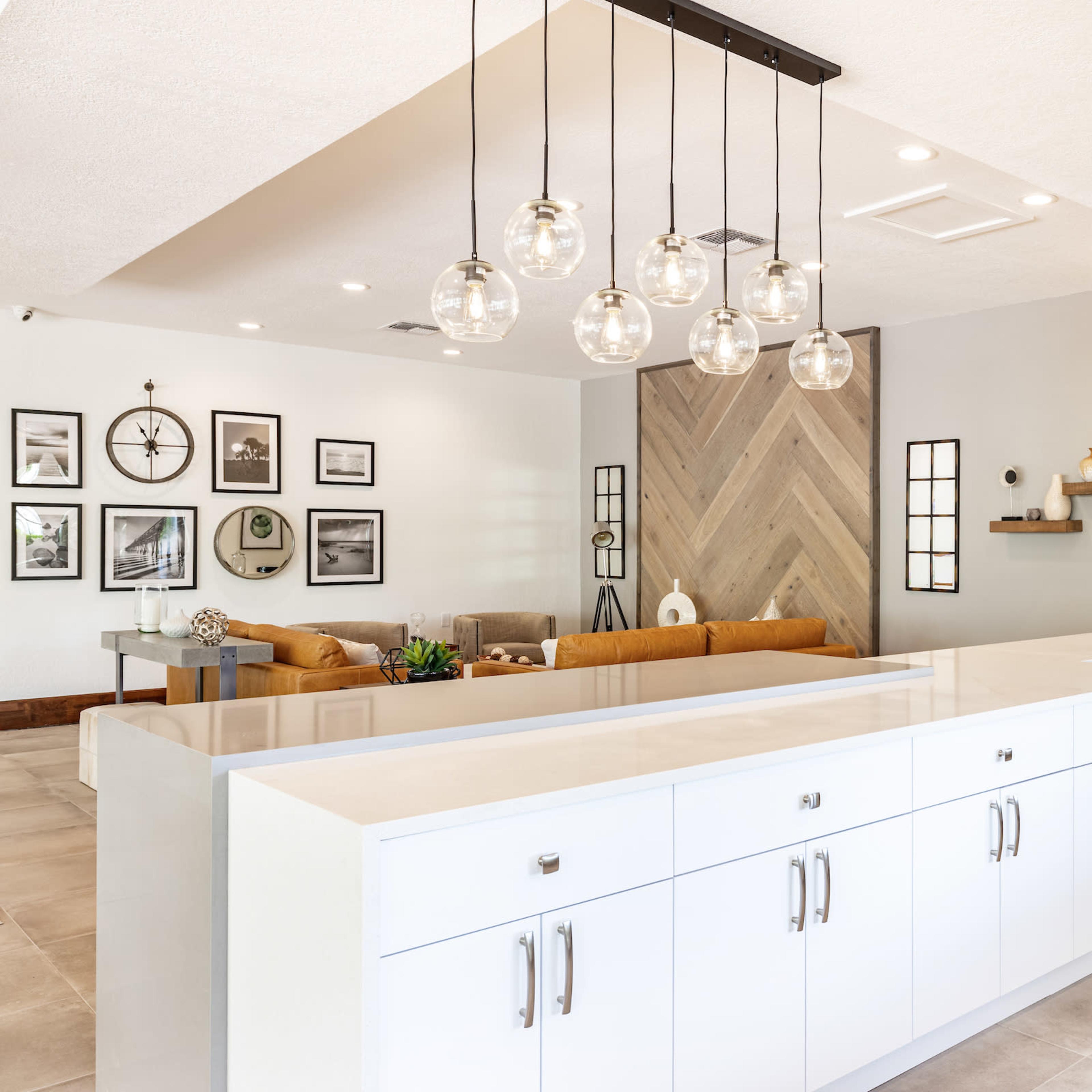 A modern kitchen with a white cabinet island, pendant lights above, and a living area visible in the background featuring framed wall art and a seating arrangement.