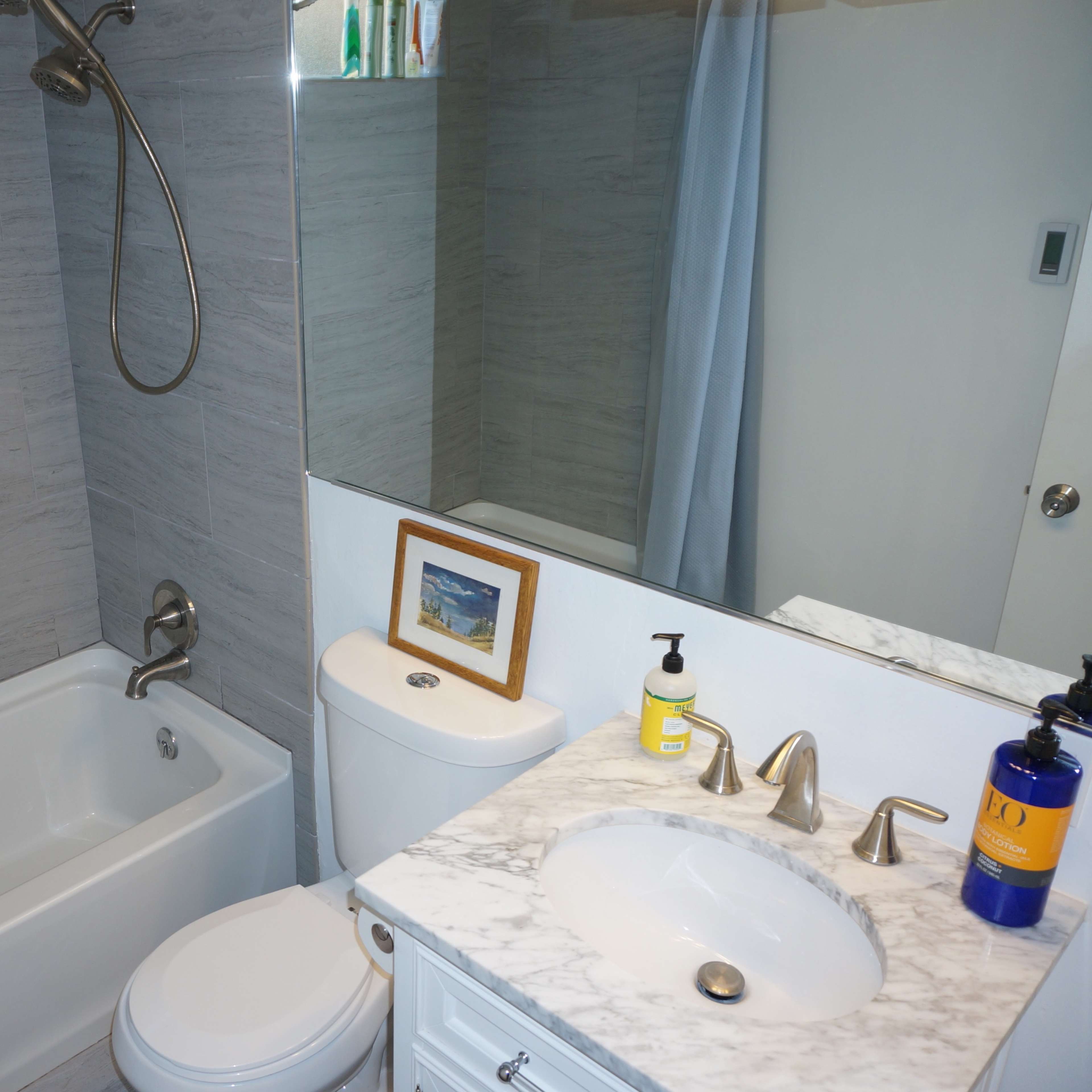 A small bathroom featuring a tub with a showerhead, a toilet, and a marble vanity with a sink and framed picture.