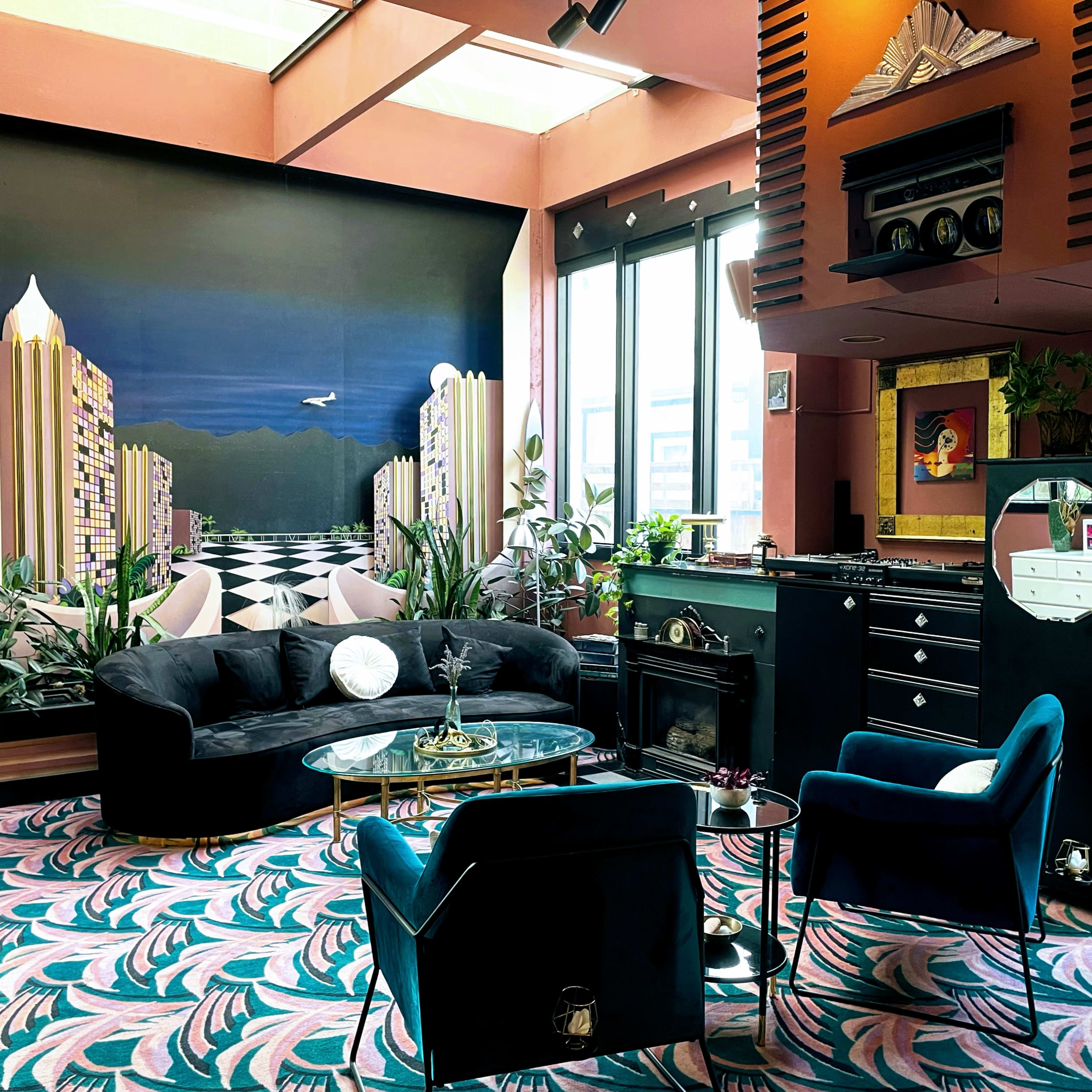 The scene features a stylish lounge area with a dark-colored couch and armchairs, surrounded by large windows and decorated with potted plants and a vibrant patterned carpet.