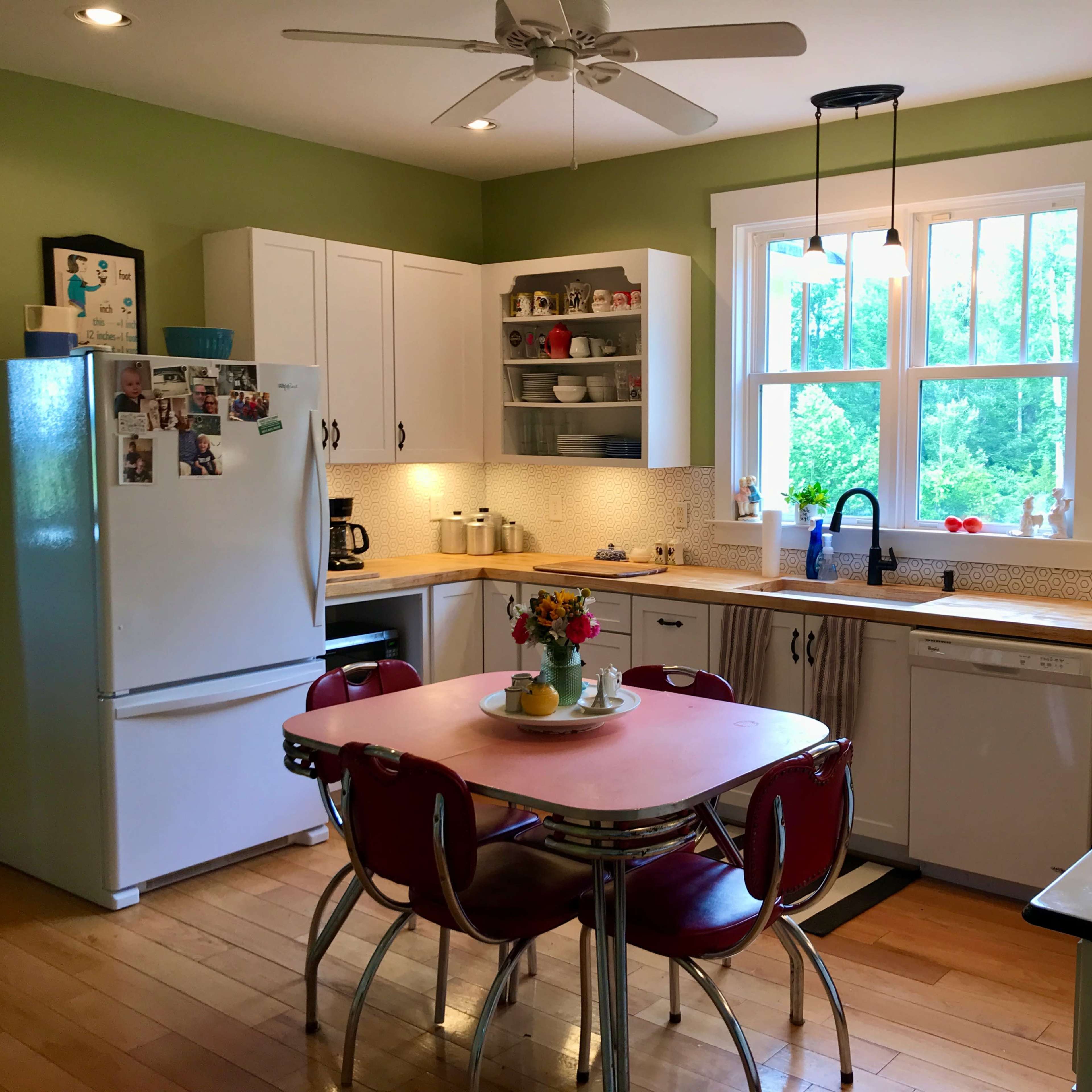 A well-lit kitchen features green walls, a central table with red chairs, and white cabinets displaying dishes and a window overlooking greenery.