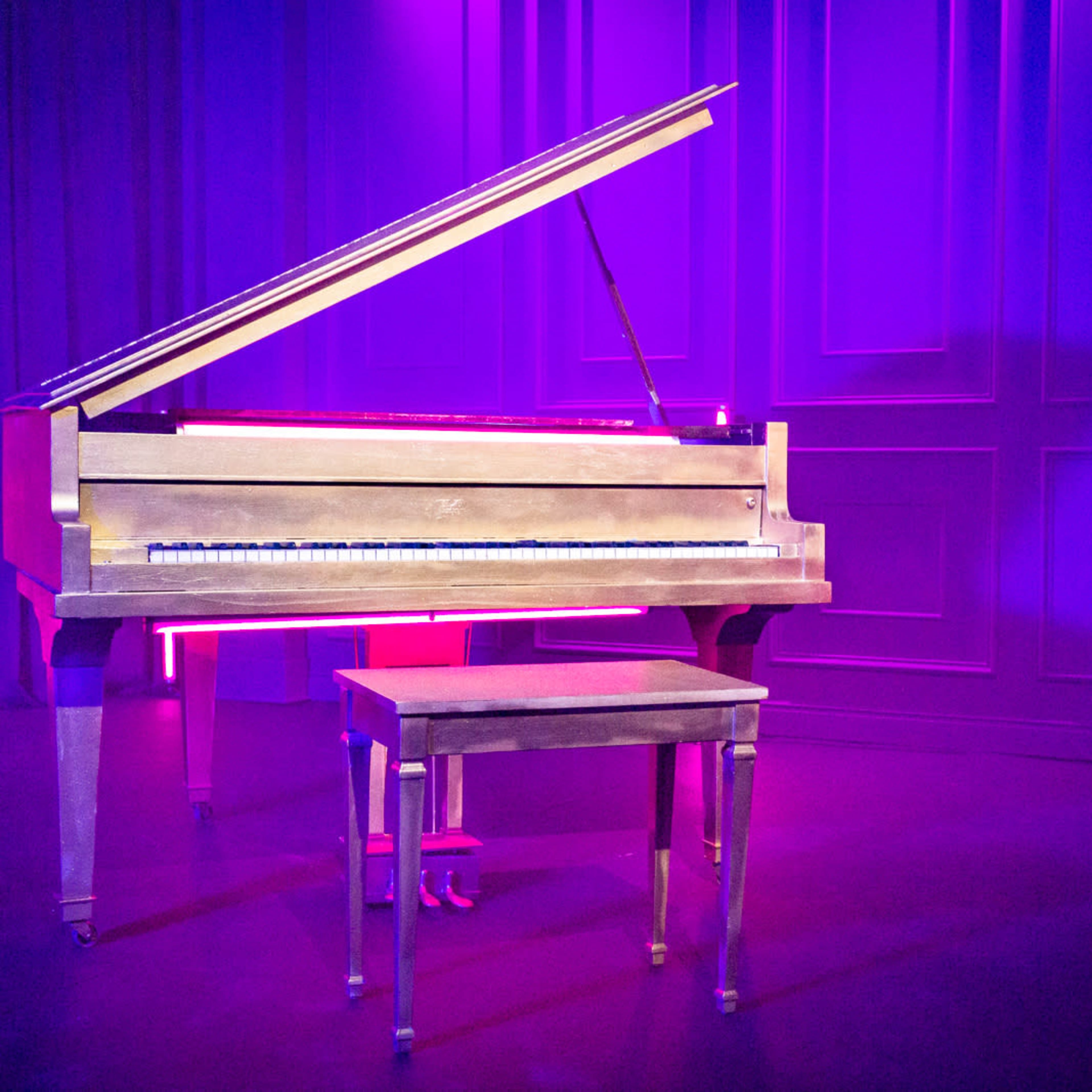 A grand piano with its lid raised sits next to a matching bench beneath purple lighting on a stage.