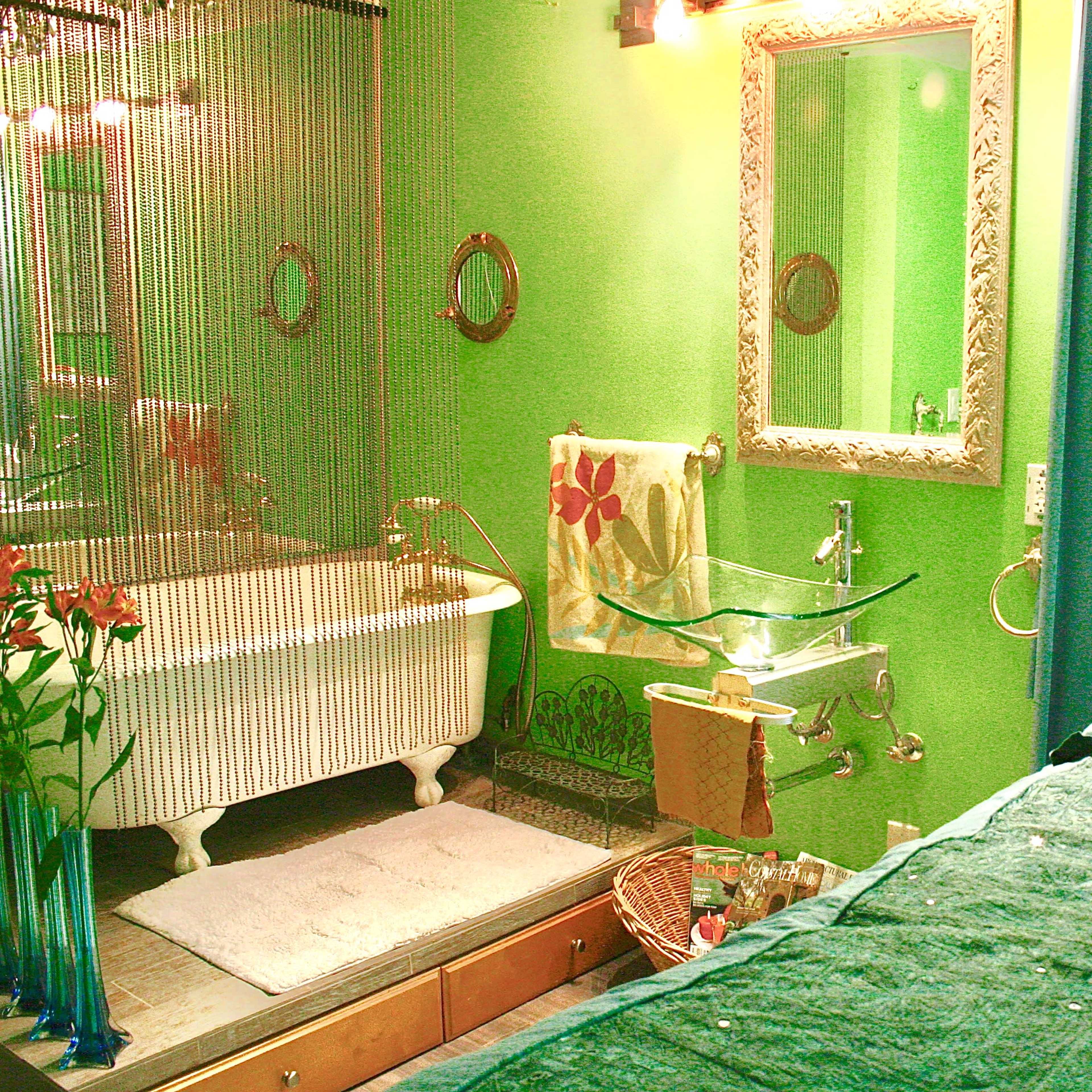 A colorful bathroom with a clawfoot tub, a glass sink vanity, and a bed draped with a blue coverlet and orange pillow.