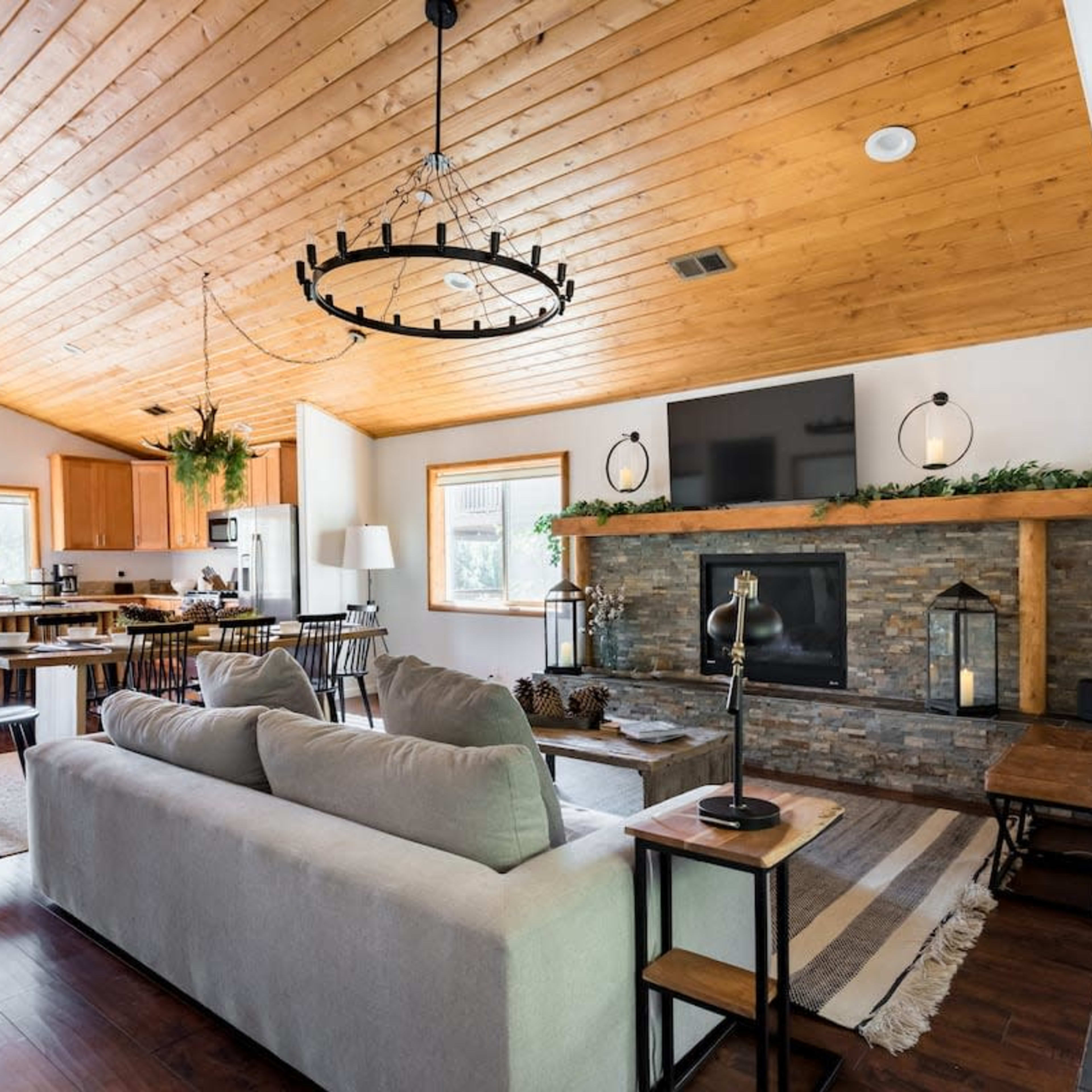 A spacious living area features a wooden ceiling, a stone fireplace, and modern furnishings, connecting to an open kitchen.