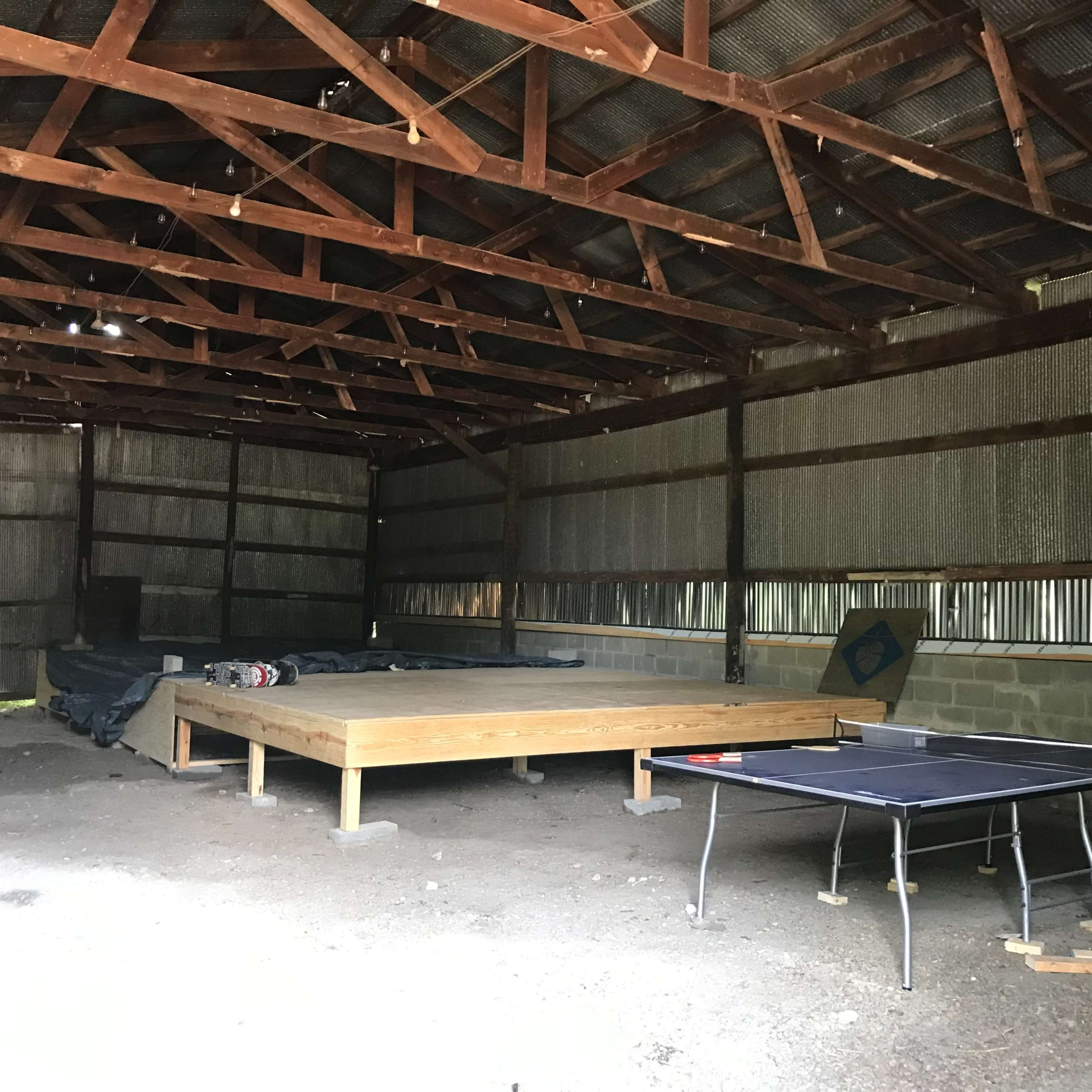 An interior of a barn with a wooden platform and a ping pong table set on a gravel floor.