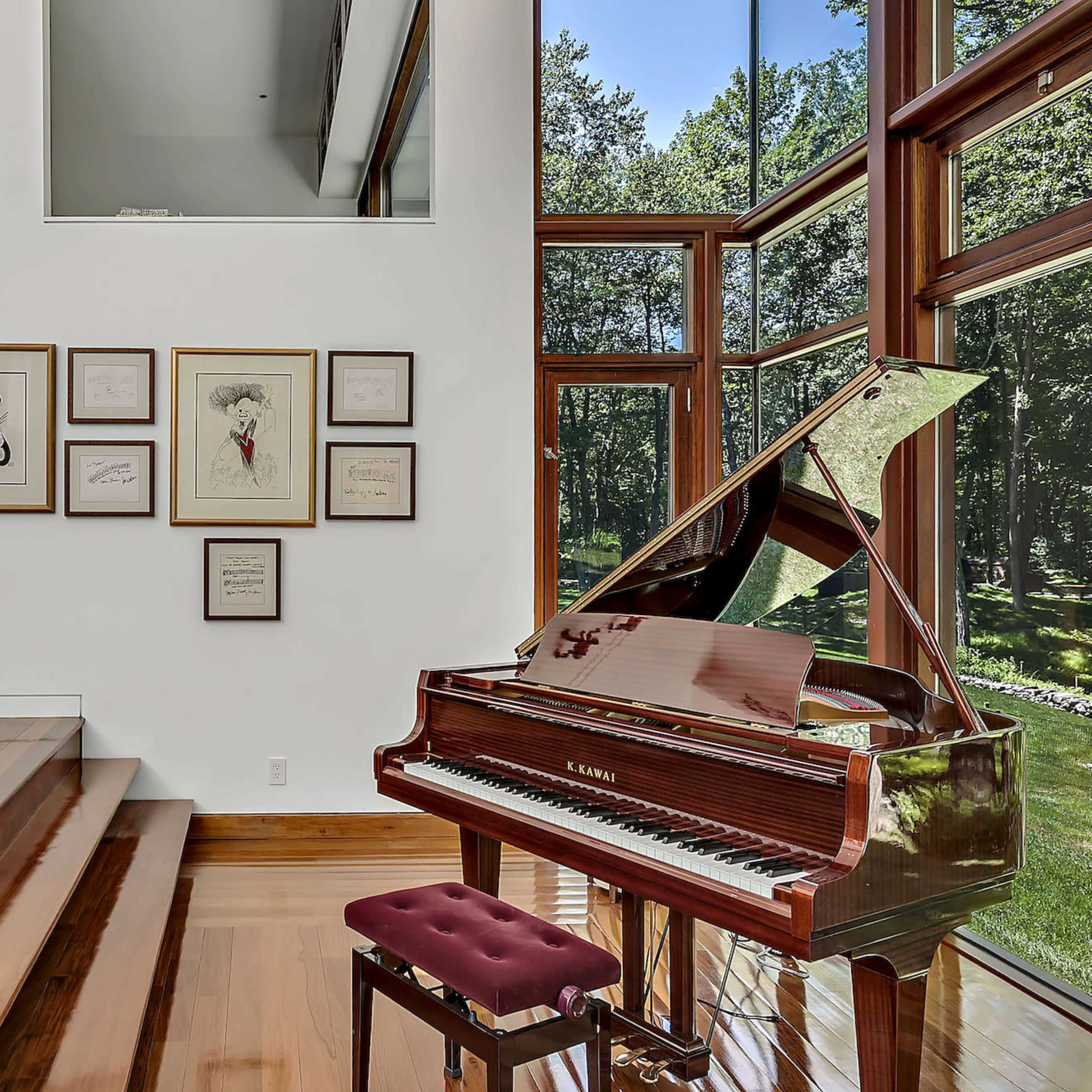 A grand piano sits beside a staircase in a room with large windows overlooking a wooded area, adorned with framed artwork on the wall.