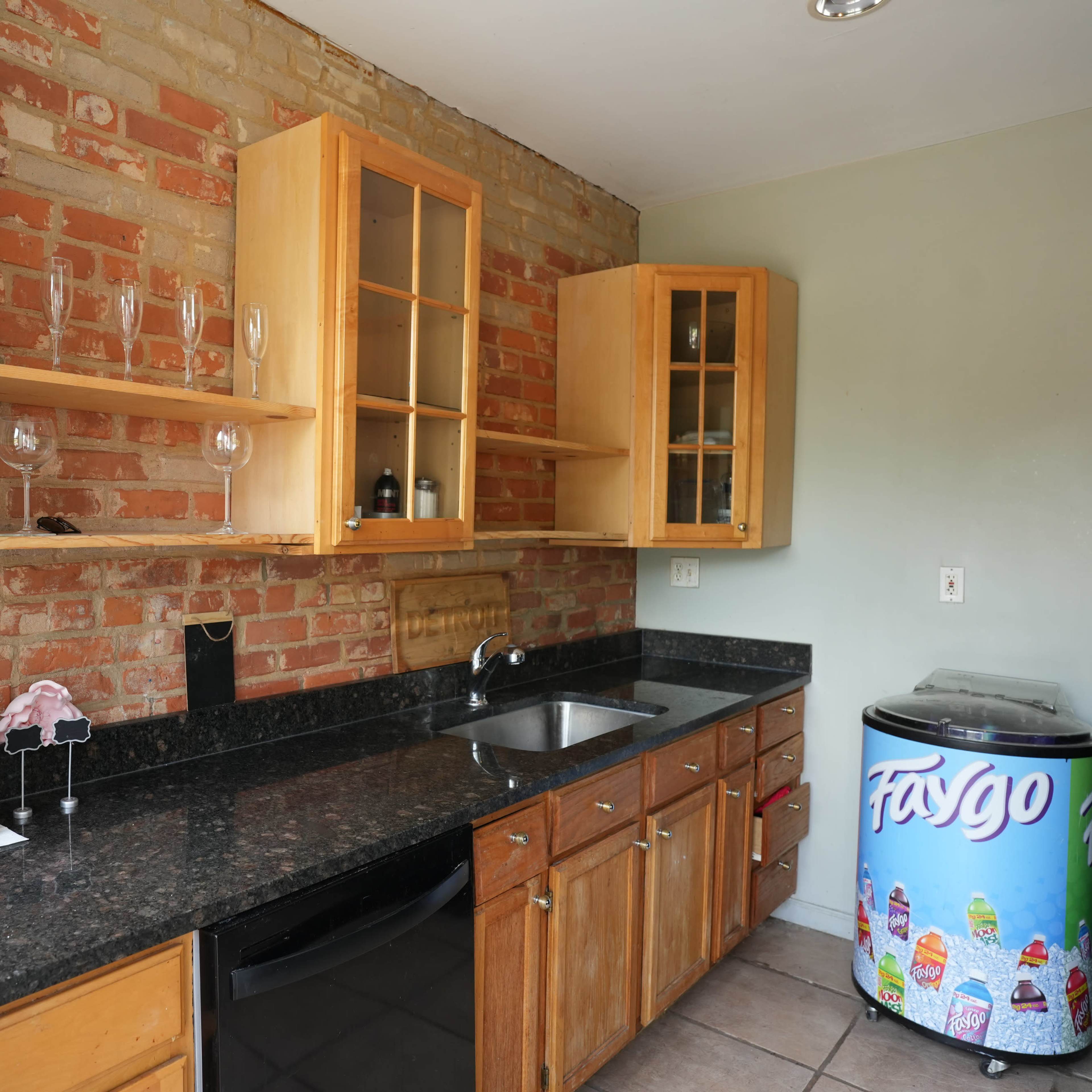A kitchen with exposed brick walls, wooden cabinets, a black countertop, a sink, and a colorful beverage fridge.