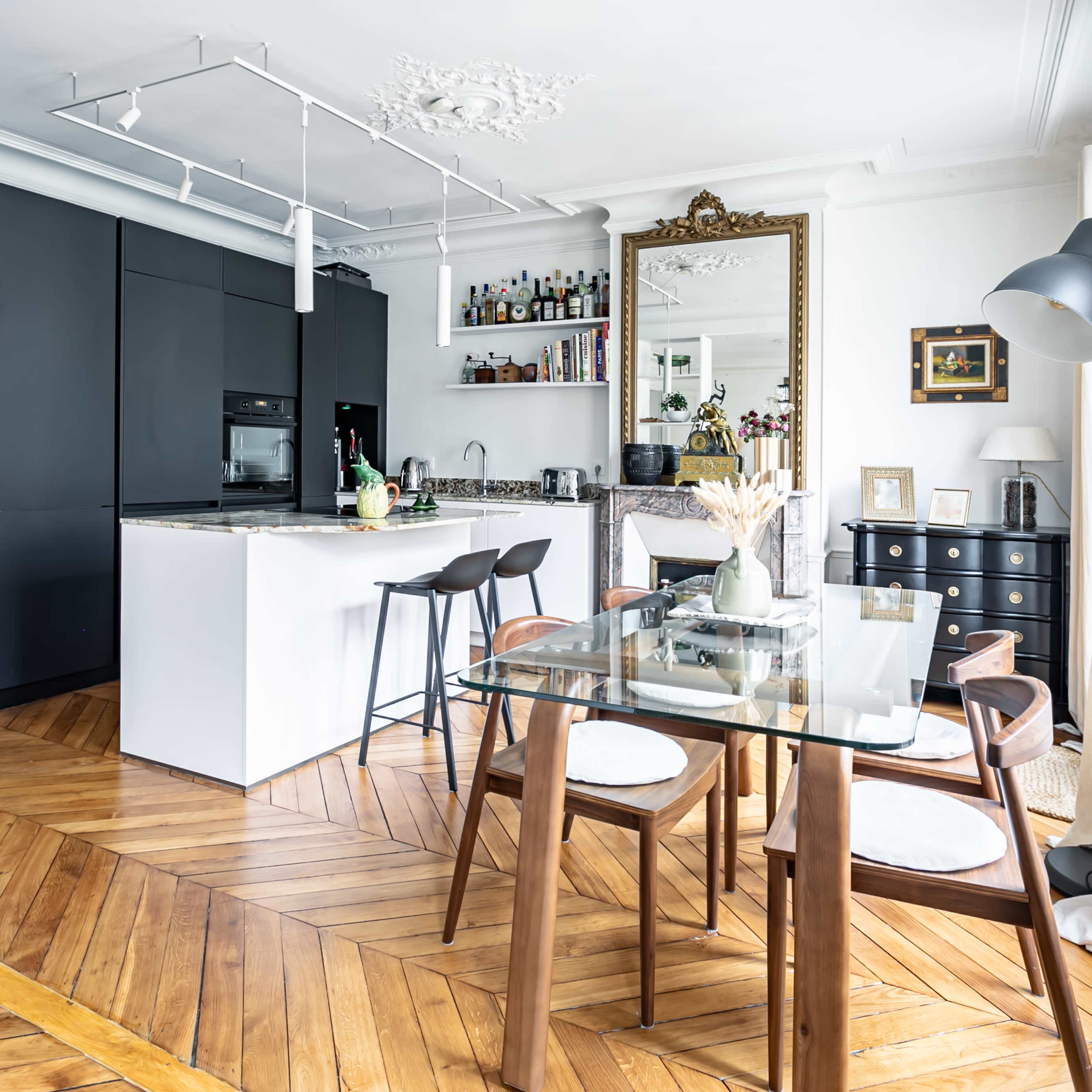 A modern kitchen and dining area features sleek black cabinetry, a glass-top dining table with wooden chairs, and a mirror reflecting the room's decor.