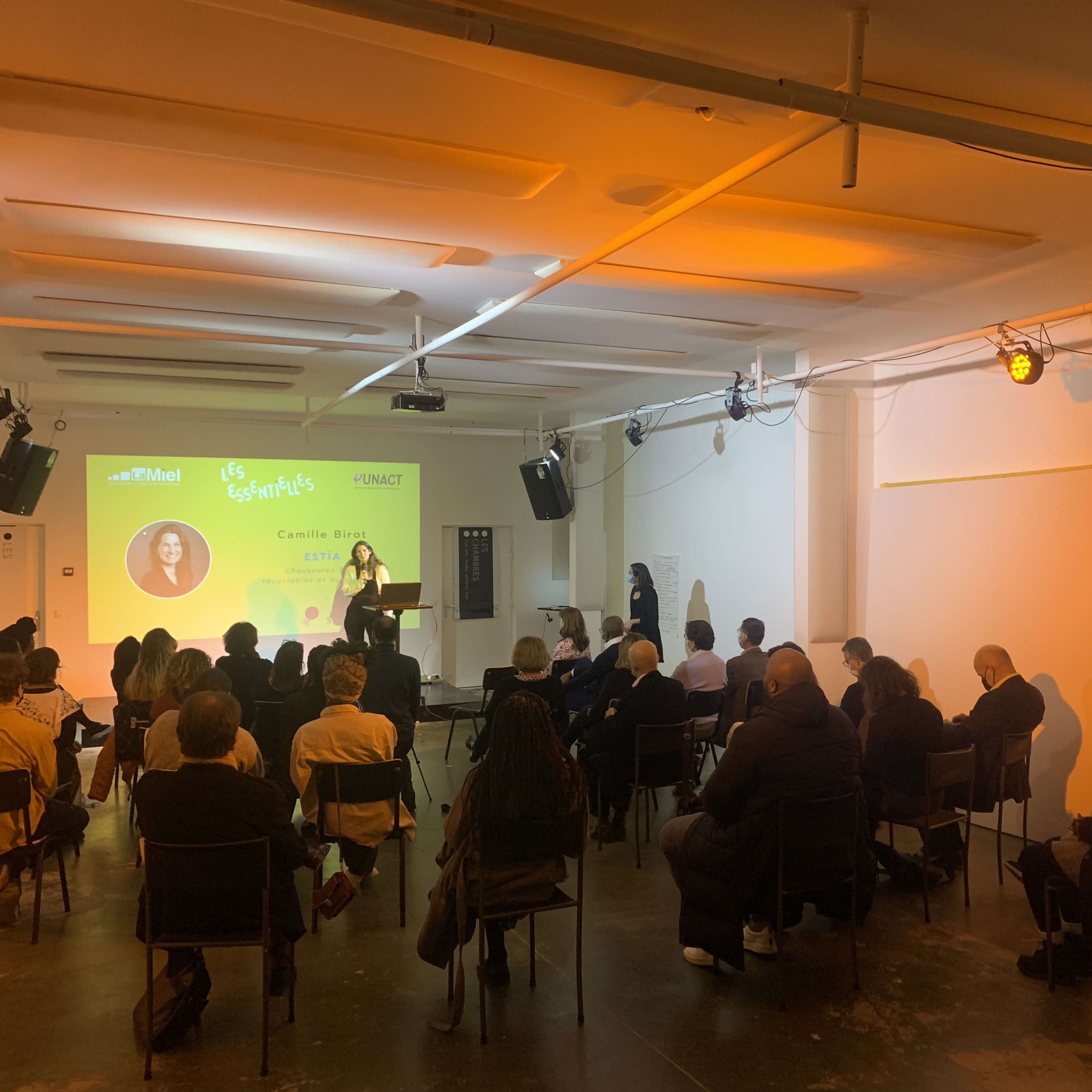 A speaker presents to an audience seated in a brightly lit room with a projector screen displaying information.