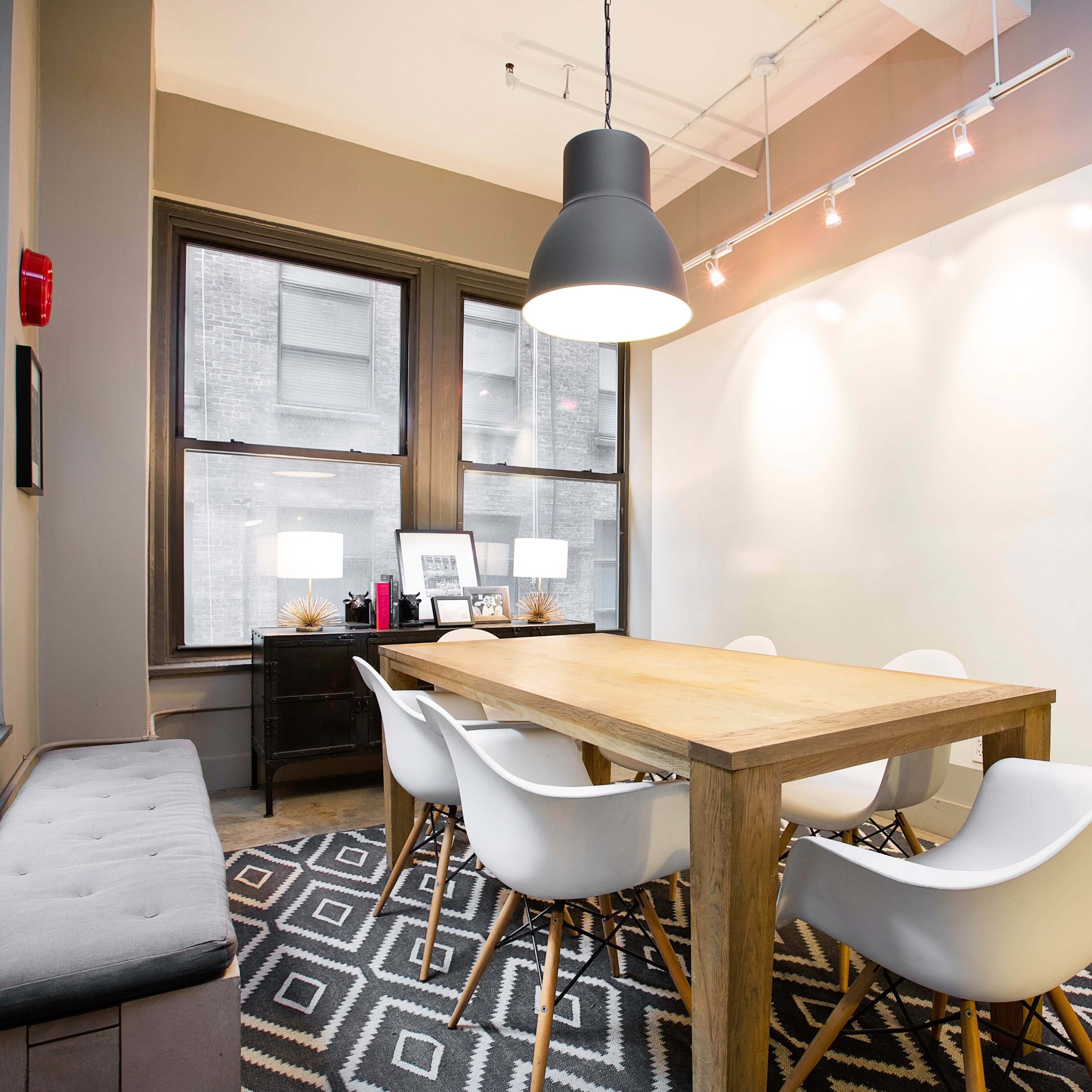 A meeting room features a large wooden table surrounded by white chairs, with windows showcasing a city view and a geometric-patterned rug on the floor.