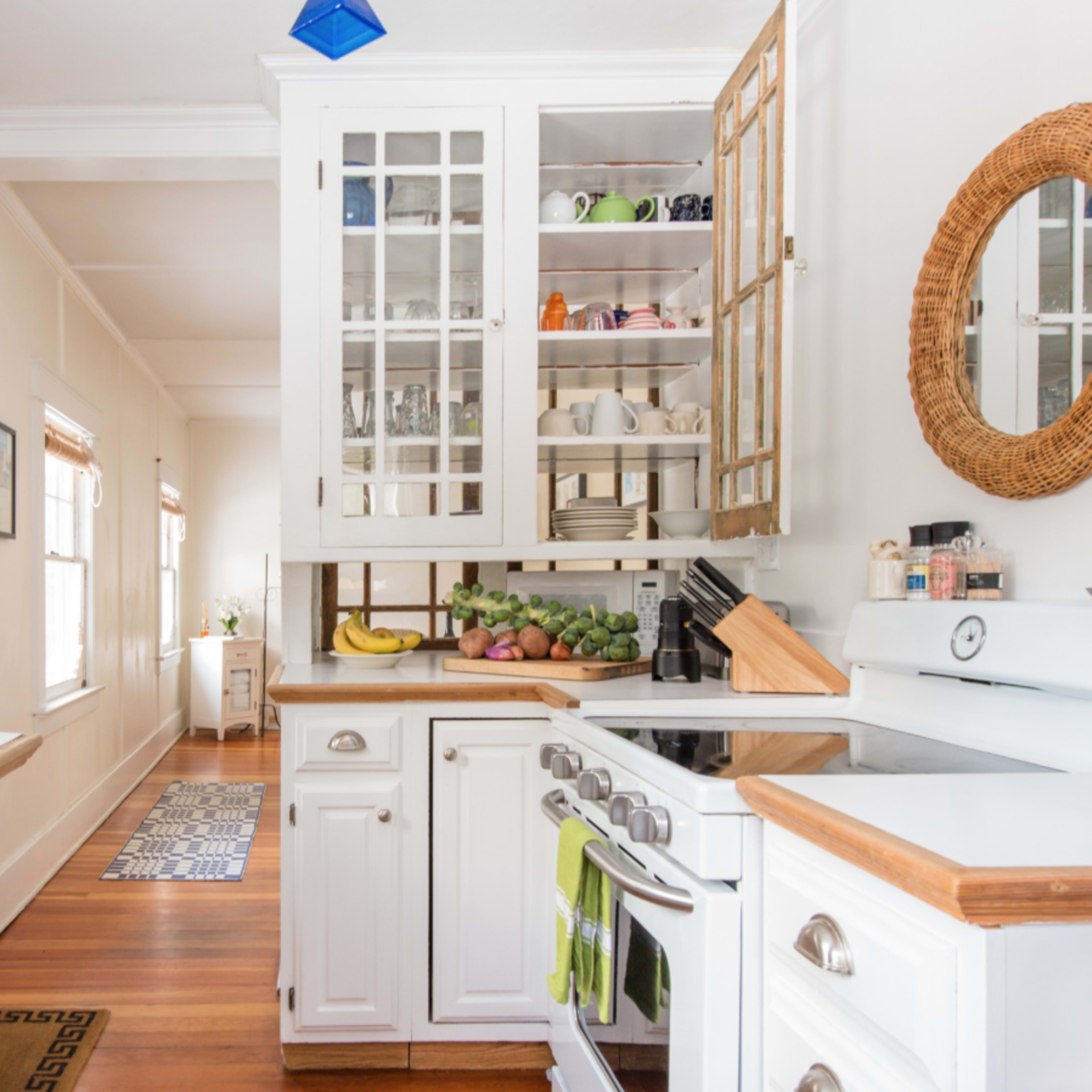 A bright kitchen features white cabinetry, a sink, a stove, and a glimpse of a hallway with a rug.